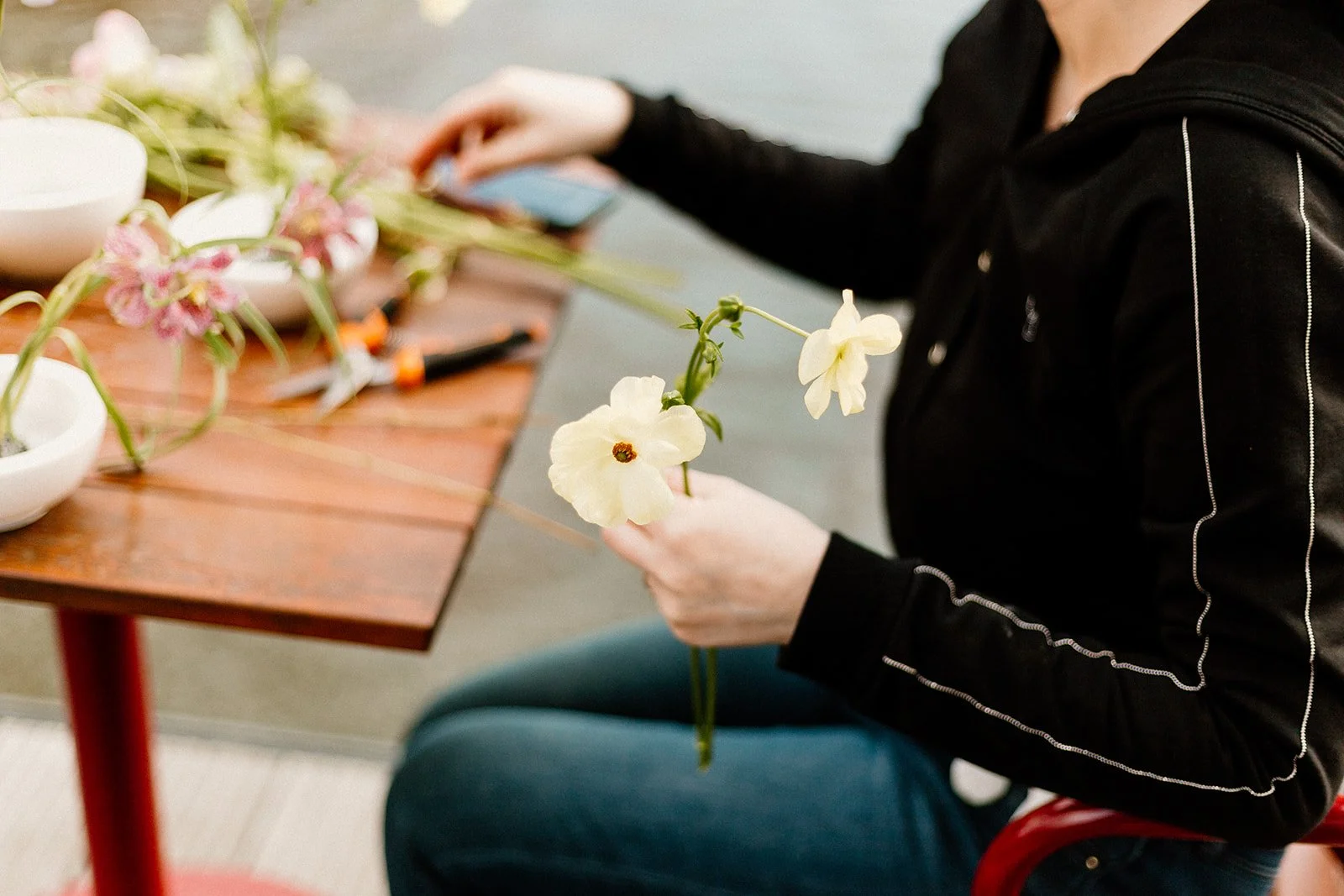 Postać kobiety w czarnej bluzie trzyma jasno żółte kwiaty w dłoni , siedzi przy stole na którym jest sekator i białe miski z kwiatami.