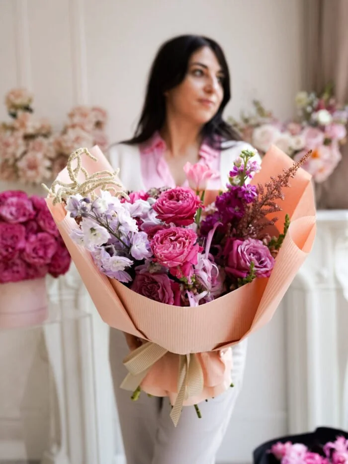 Kobieta trzymająca duży bukiet różowych i fioletowych kwiatów otoczonych różowym papierem do pakowania, stojąca w pokoju udekorowanym różnymi kompozycjami kwiatowymi.