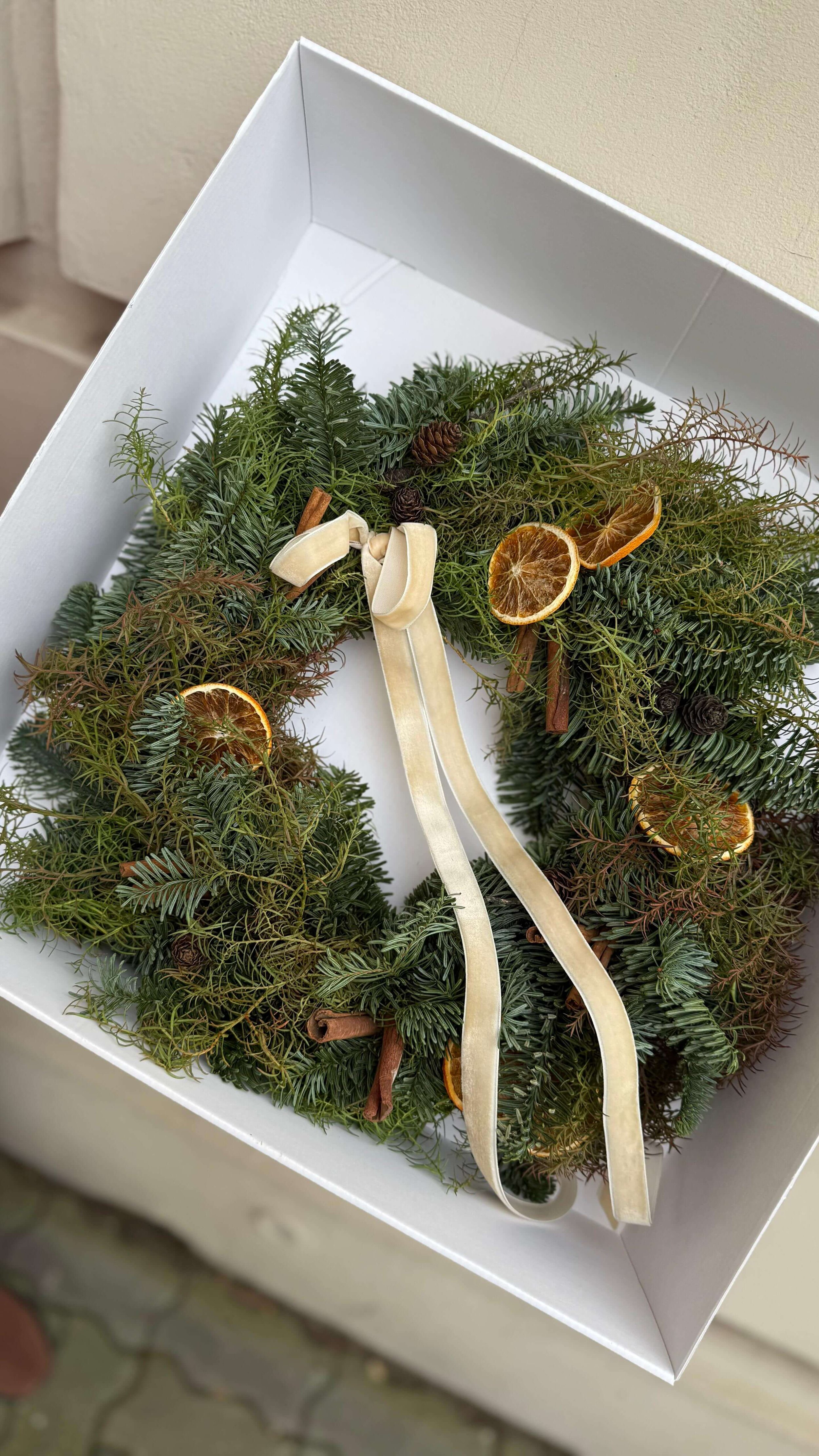 Świąteczny zielony wianek z cyprysu i jodły nobilis leżący w białym pudełku, ozdobiony małymi szyszkami,suszonymi plastrami pomarańczy i cynamonem oraz beżową welurową cienką wstążką.