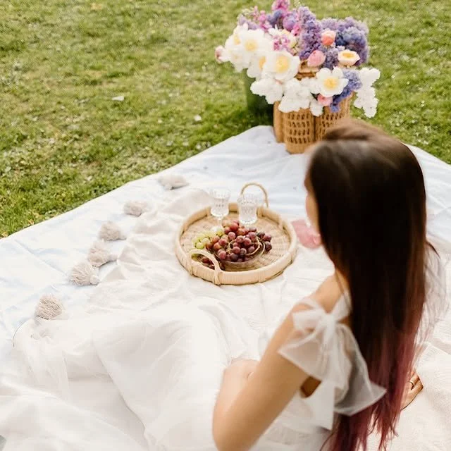 Piknik na białym kocu z wiklinową tacą z winogronami i dwiema szklankami wody, duży bukiet fioletowych, białych i różowych kwiatów w wiklinowym koszu oraz kobieta z długimi brązowymi włosami siedząca na kocu na trawie na świeżym powie