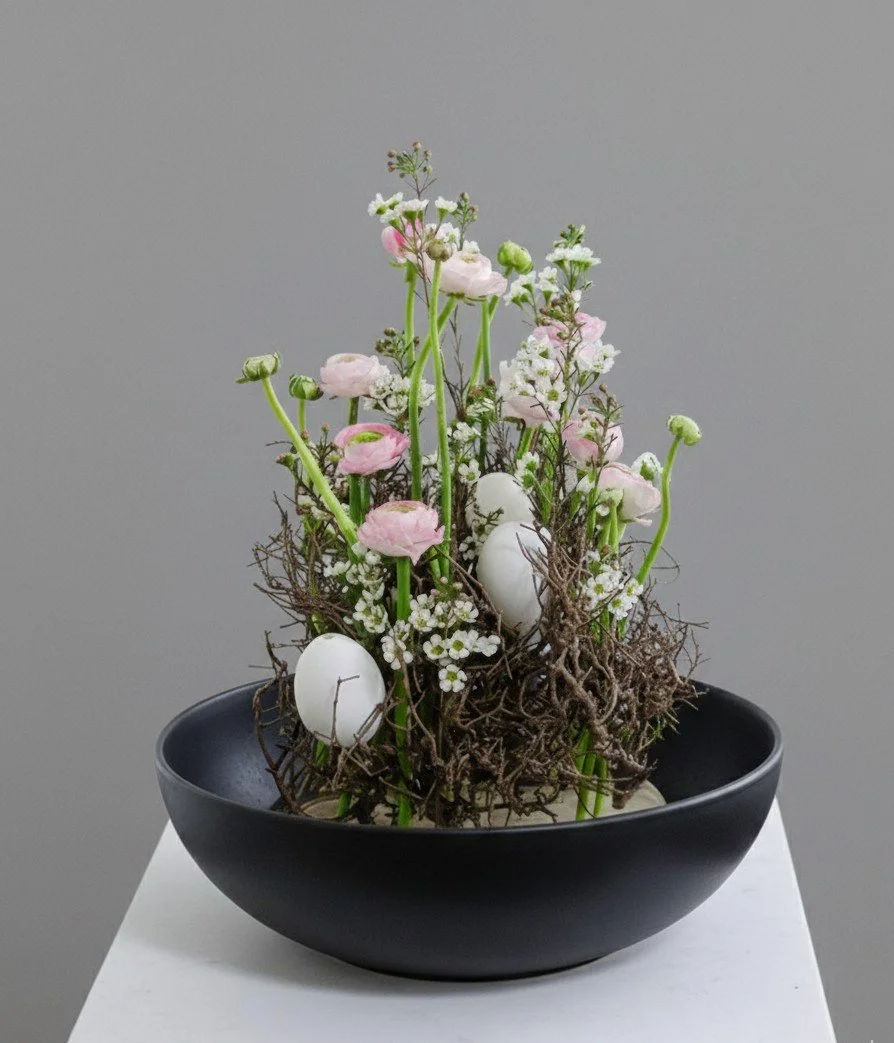Dekoracyjna kompozycja roślinna w czarnej misie z różowymi i białymi kwiatami, białymi jajkami, gałązkami i mchem.