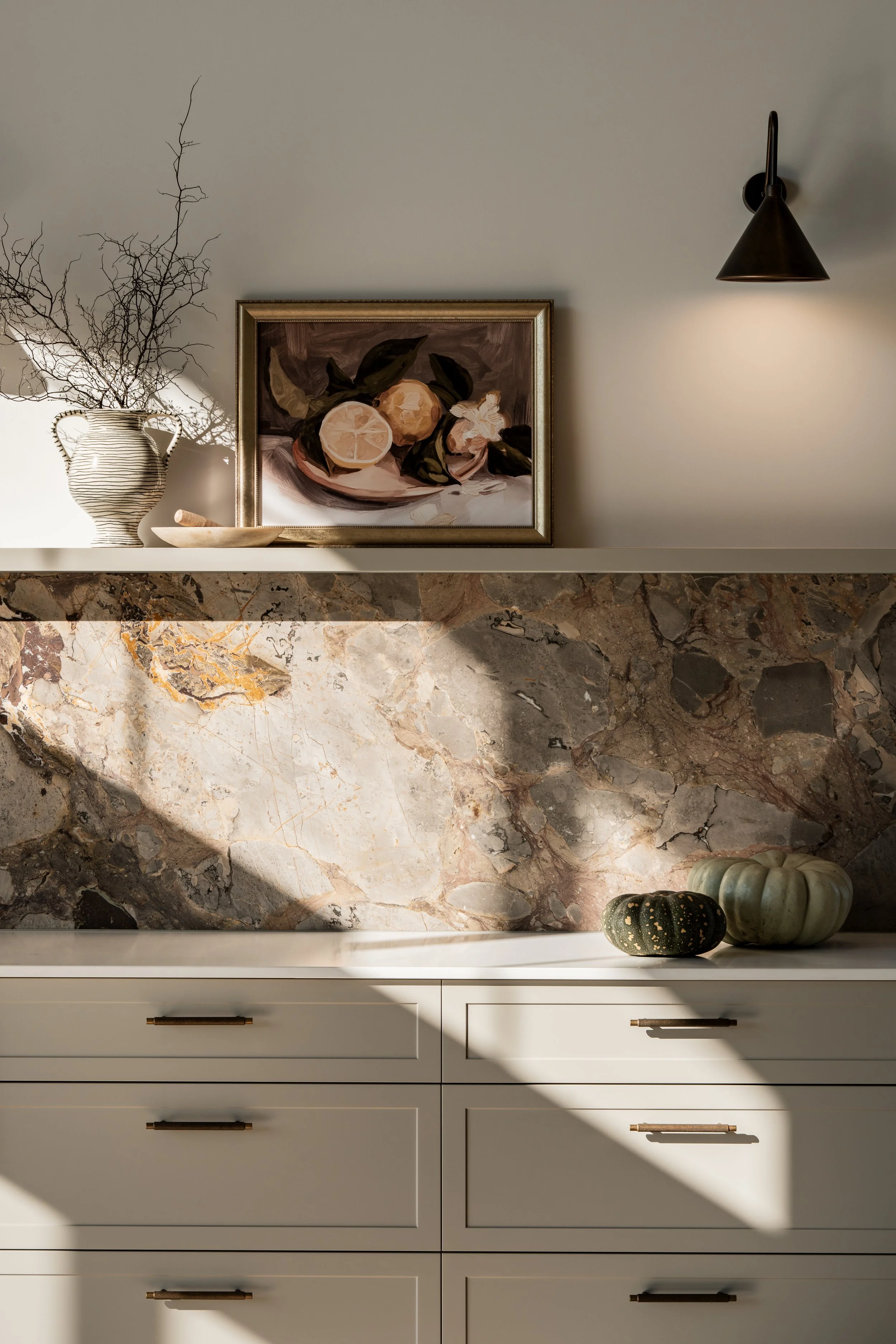 Styled kitchen shelf with framed artwork, stone backsplash, sculptural objects and directional sunlight