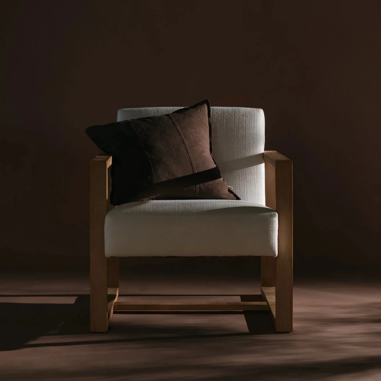 Shot by Swell Studios for Thread Haus Co. A white armchair with light wooden frame against a plain dark brown wall, decorated with a dark brown pillow.