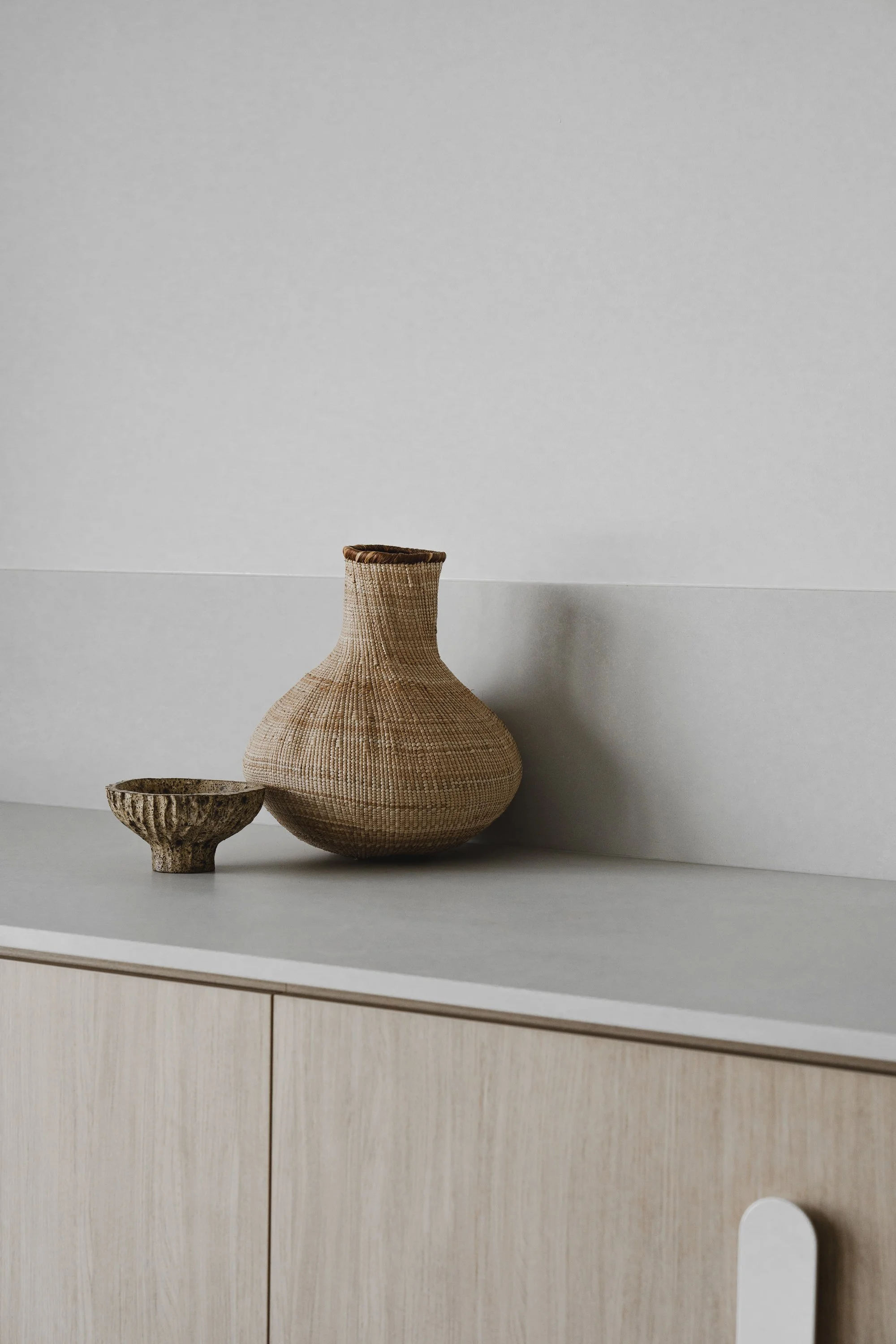 A large, rounded wicker basket-shaped vase with a narrow opening at the top, placed next to a smaller, textured bowl on a white shelf against a plain light gray wall.