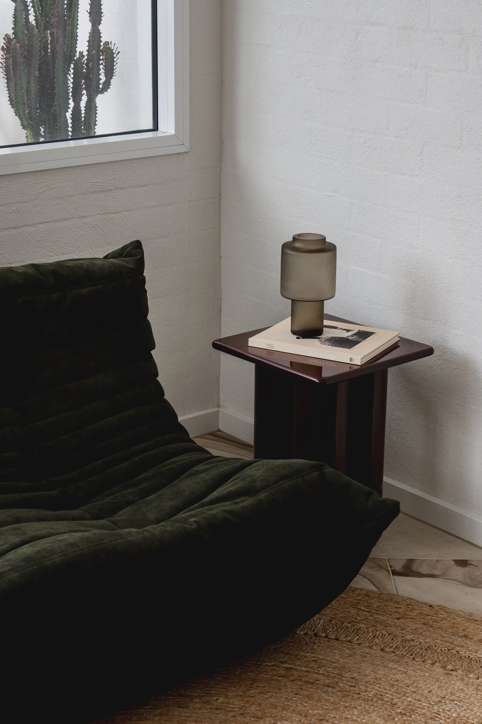 A green velvet couch in a room with wooden flooring, a window with a cactus outside, a dark wooden side table with a frosted glass lamp and books, and a woven rug.