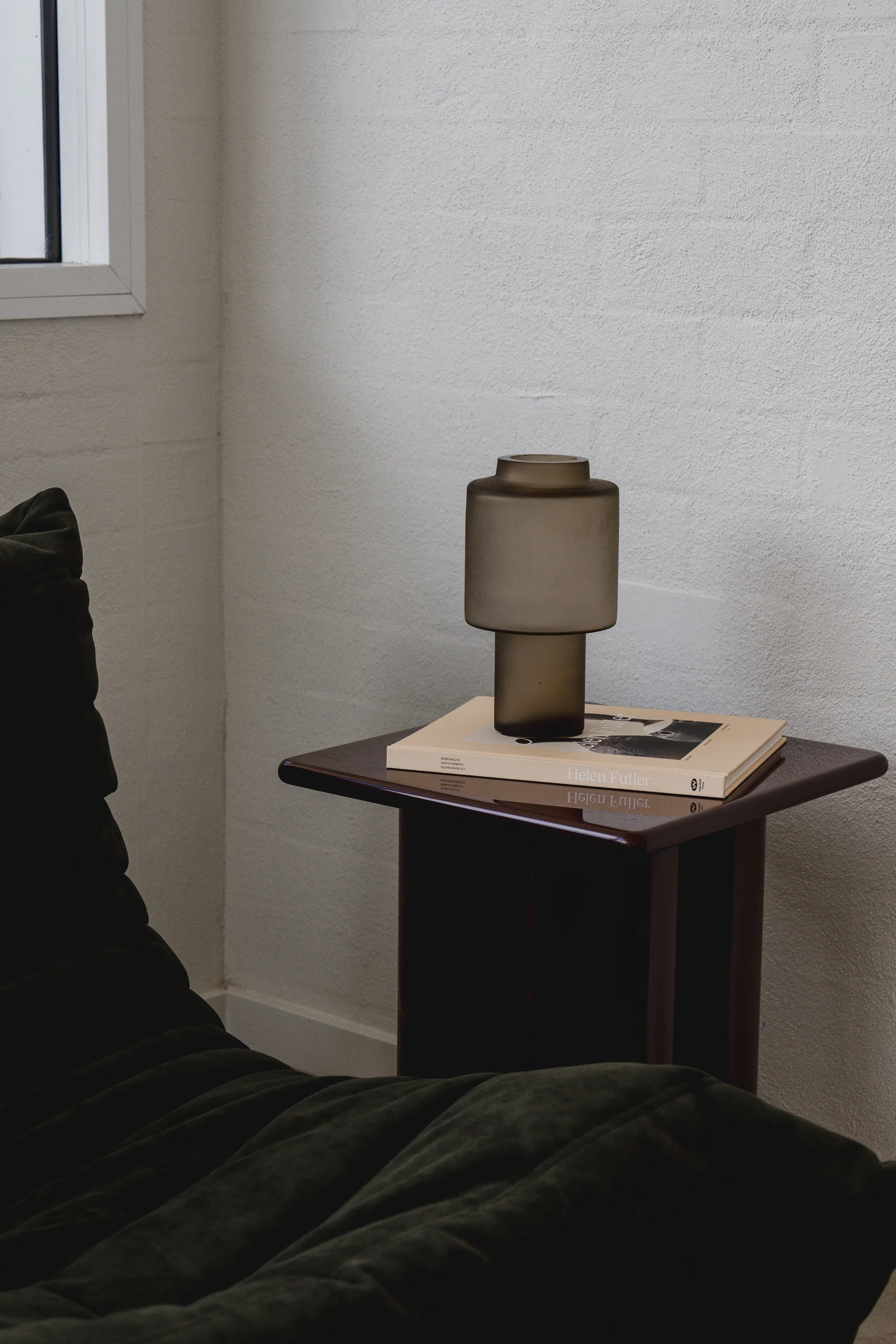 View of a small wooden table with a beige books, titled 'Helen Fuller,' and a decorative frosted glass vase on top. The table is against a white textured wall in a room with a corner window visible on the left.