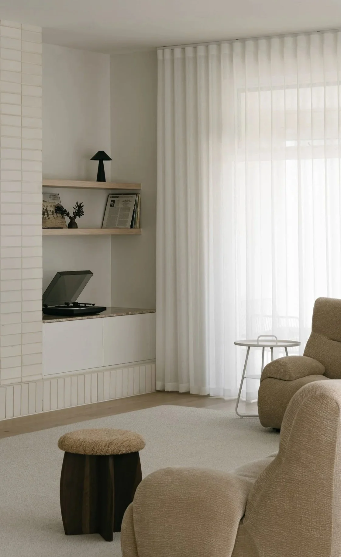 Living room with beige armchair, small round side table, beige carpet, white curtains, built-in shelf with black lamp, vinyl record player, and framed photos, and a beige upholstered stool.