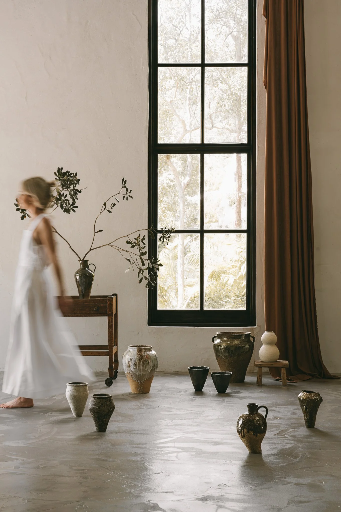 A woman in a white dress walking through a room with pottery and a large window with brown curtains.