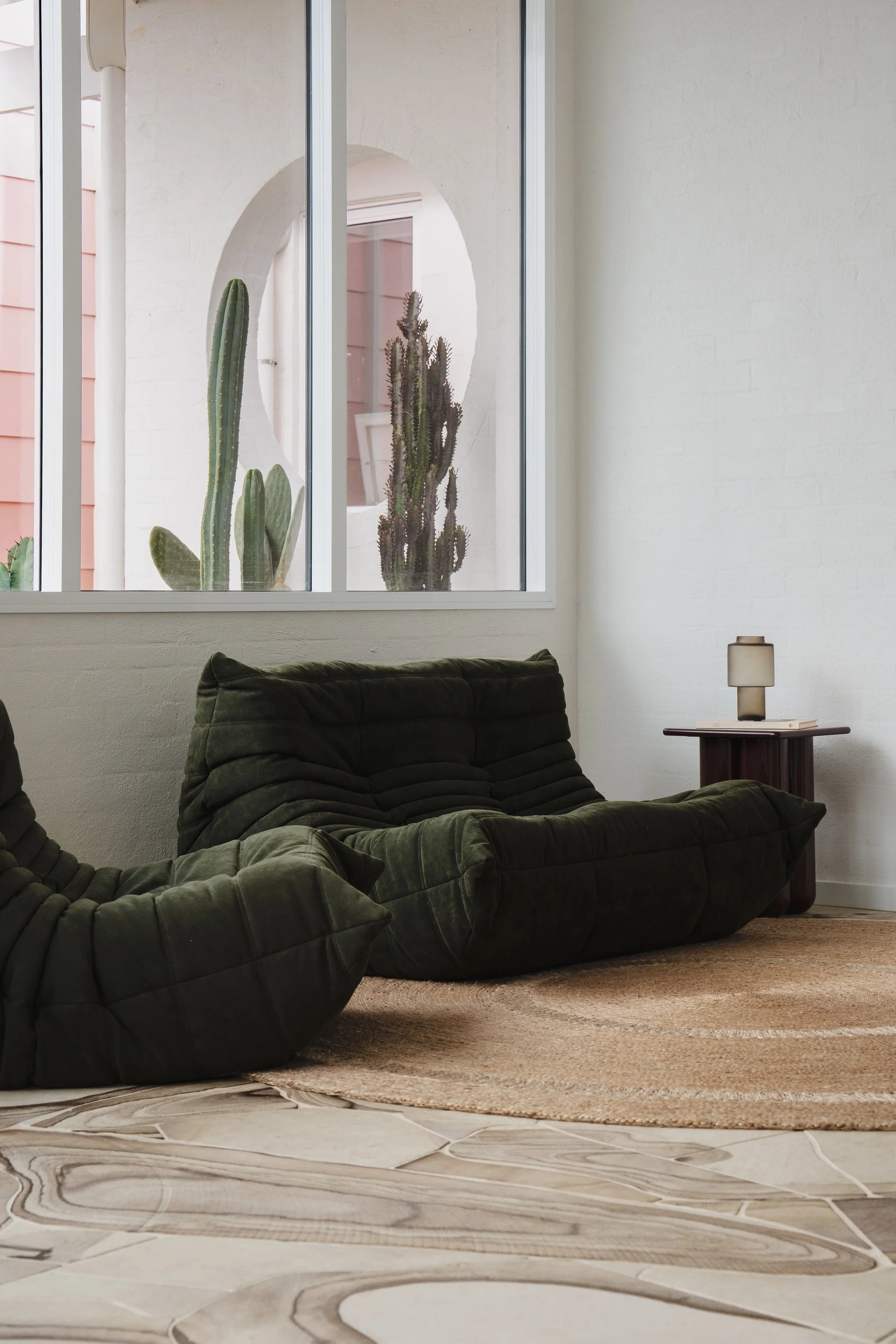 Living room with green velvet sofa, tan and white patterned rug, side table with a lamp, and a window with cactus plants outside.