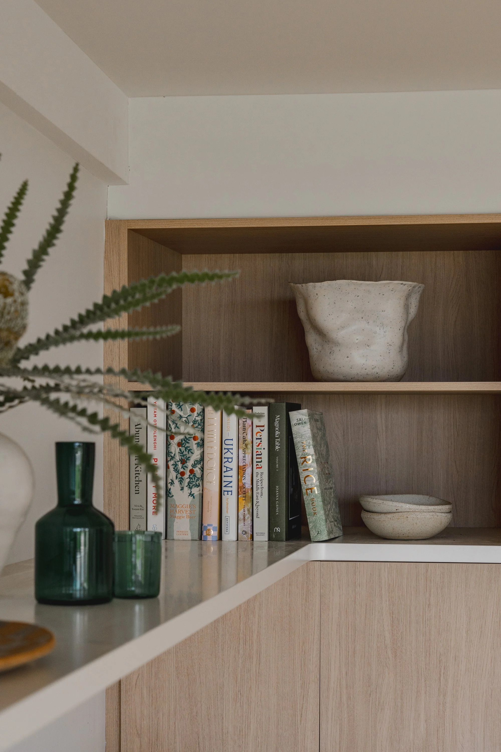 A wooden bookshelf with books, a large ceramic bowl, a small ceramic bowl, a green glass vase, and a green glass bottle with a plant in it.