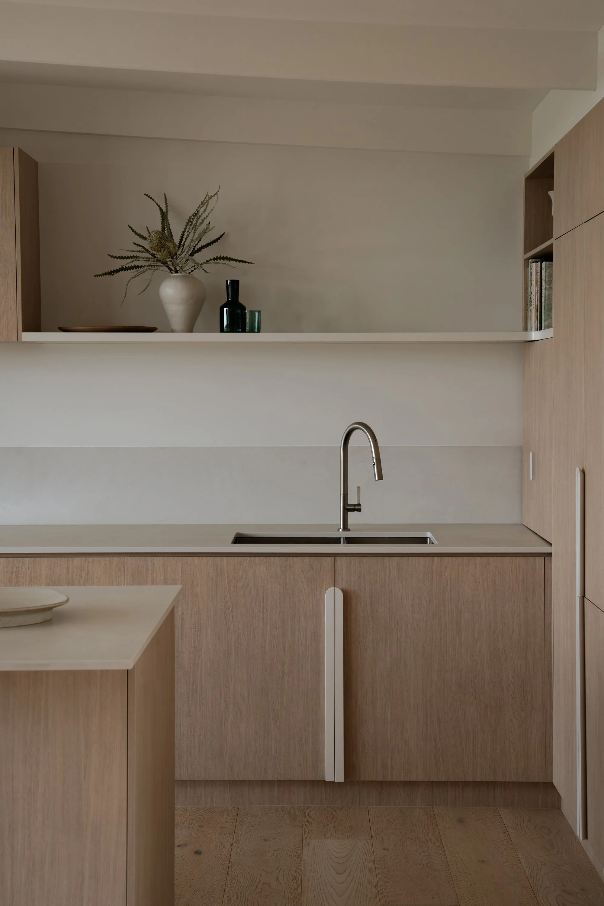 A modern kitchen with light wood cabinets, a white countertop, a stainless steel sink, and a faucet. On the shelf above, there is a white vase with dried plants and decorative bottles. The flooring is light wood.