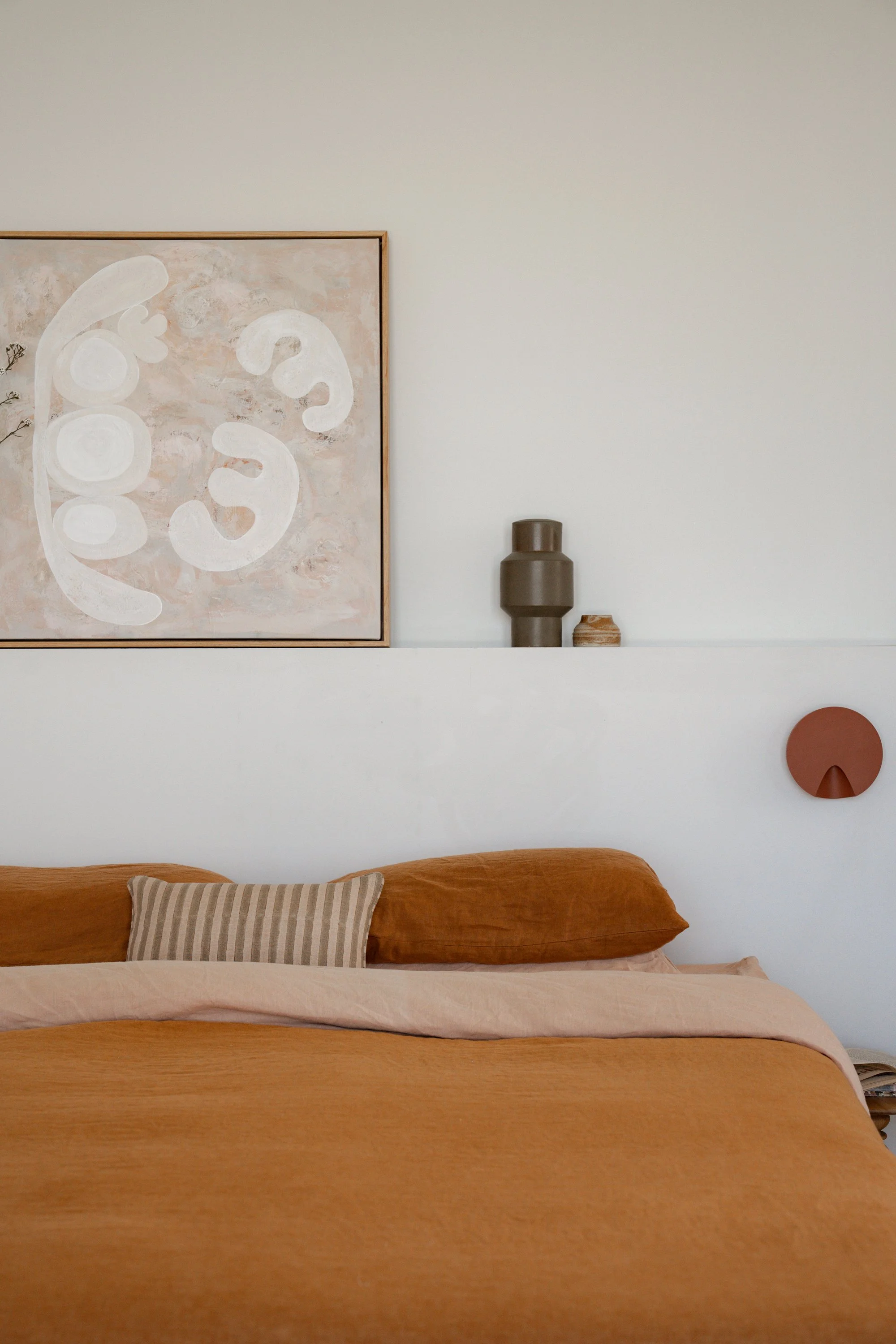 A neatly made bed with a rust-colored blanket, a light pink sheet, and a striped pillow. Behind the bed, a white wall is decorated with abstract artwork, a tall gray vase, a small bowl, and a round, rust-colored wall art piece.