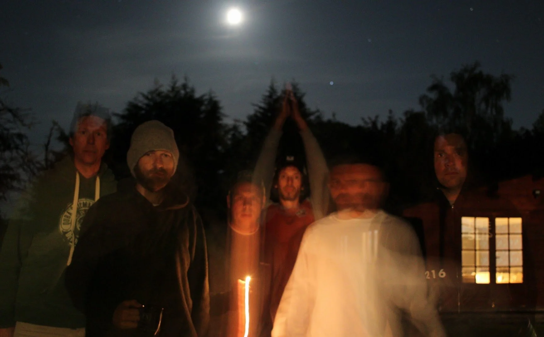 Group of men in a dark outdoor setting with blurred motion effects, holding a light source, trees and a building in the background, moon visible in the sky.