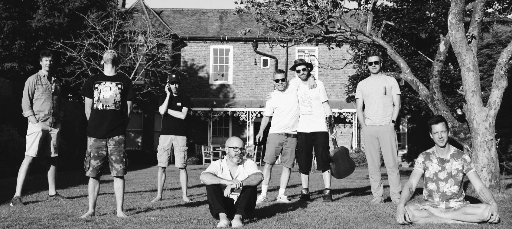 Group of eight men posing outdoors, some standing and some sitting, in front of a large brick house and trees, in black and white.