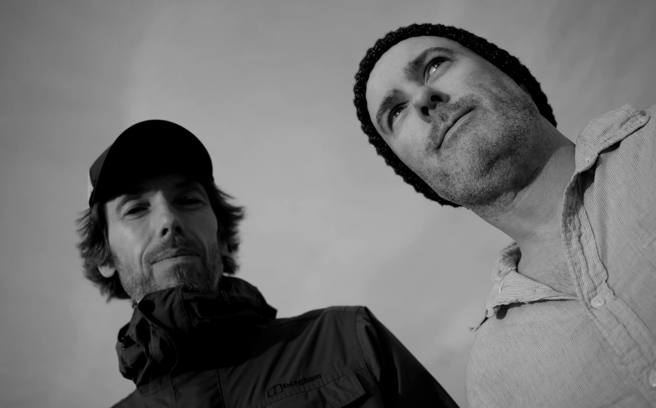 Black and white image of two men, one wearing a cap and jacket, the other a beanie and shirt, looking upwards.