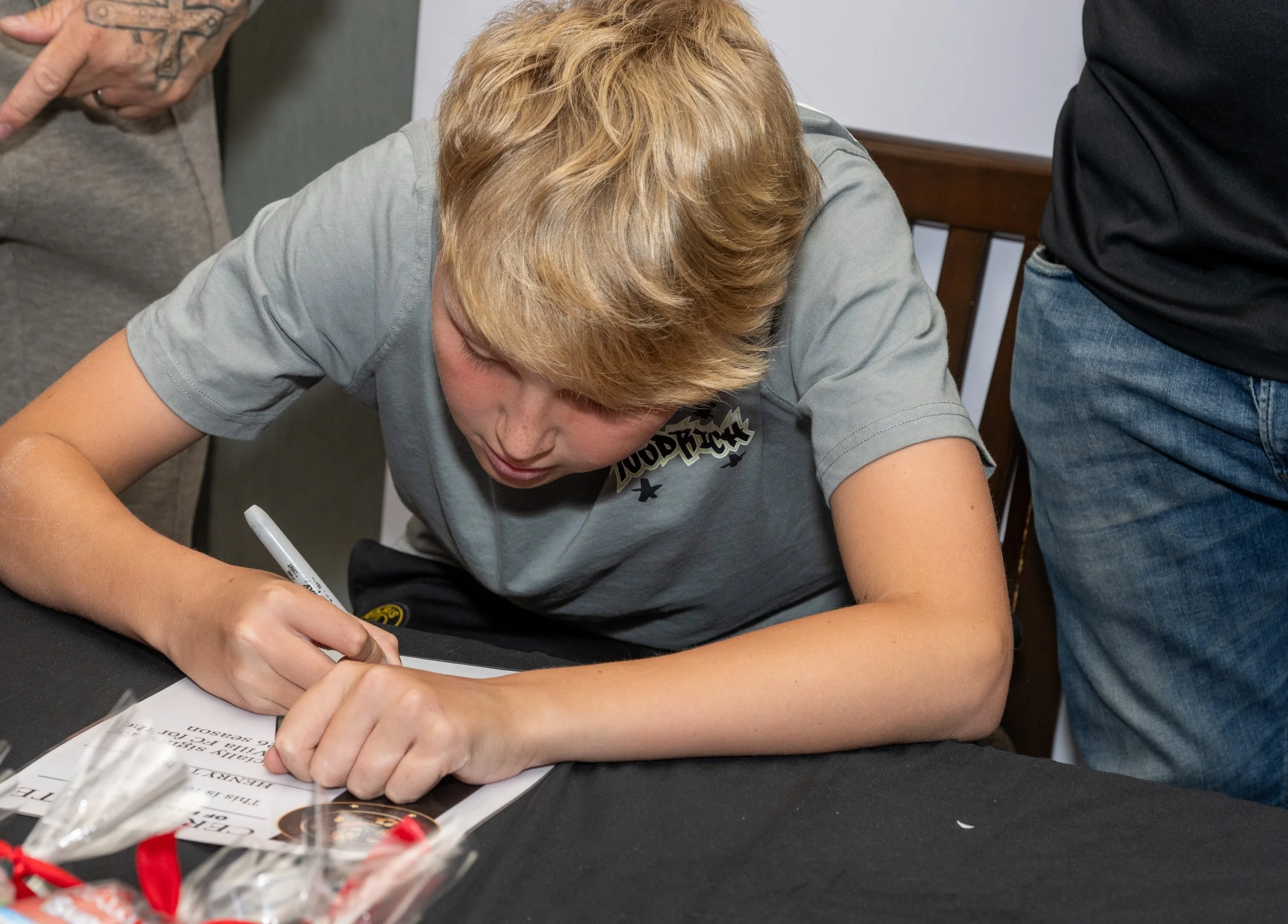 A young boy with blond hair signing a document or poster on a table surrounded by adults.