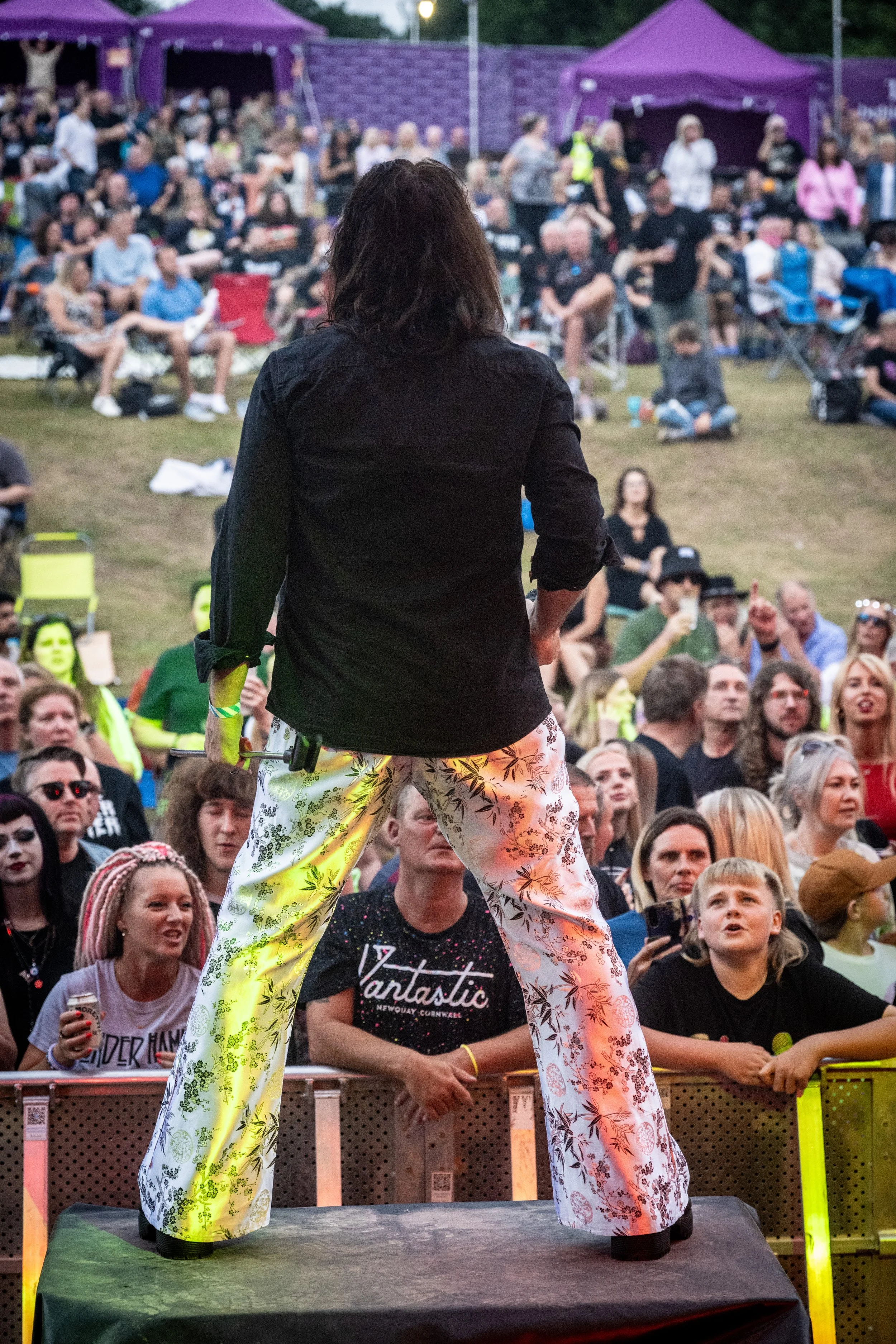 Performer on stage in front of a crowd at outdoor event with purple tents in background.