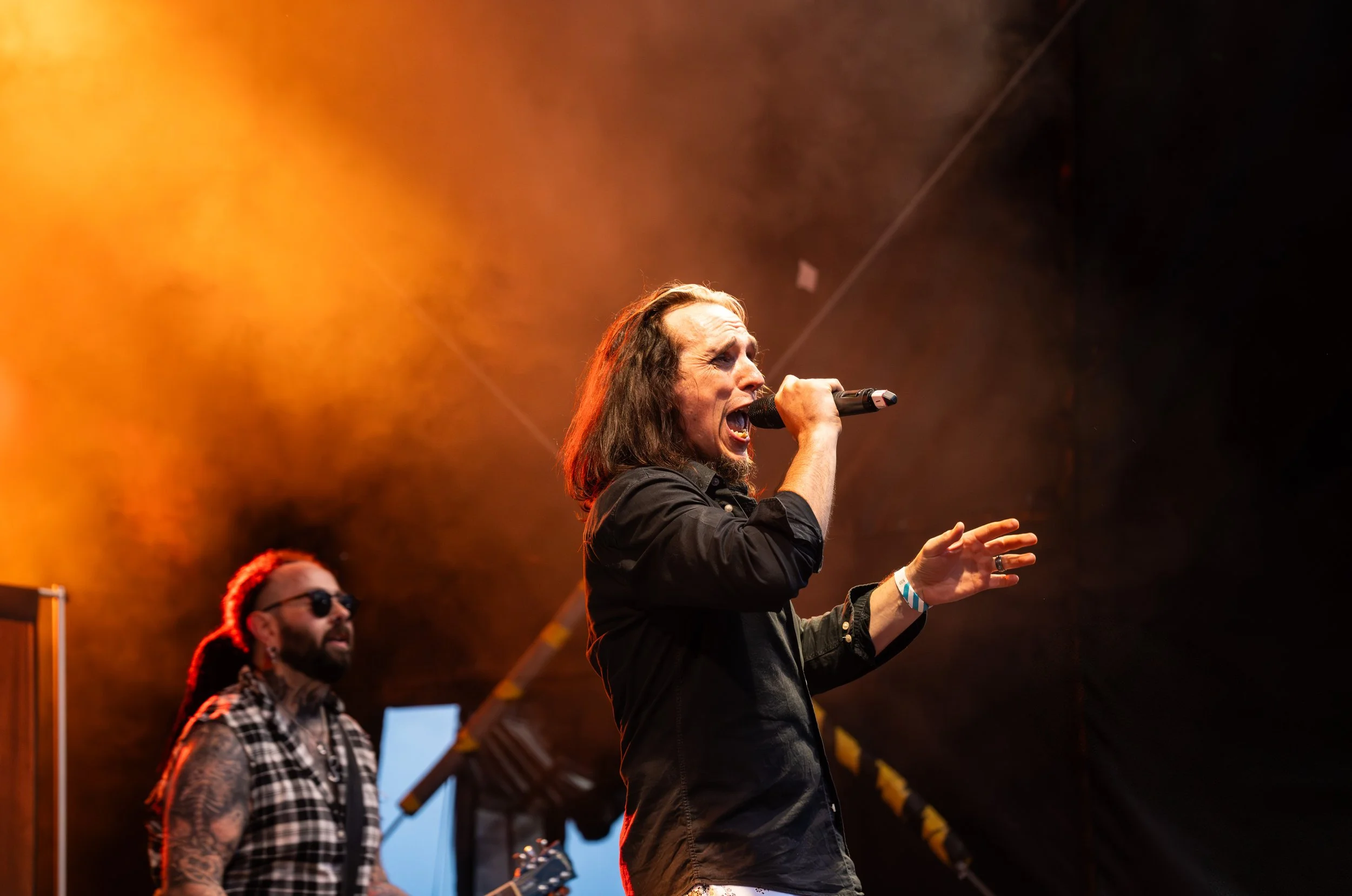 Male singer with long hair and black shirt singing into a microphone on stage during a concert, with a guitarist in the background under warm orange stage lighting.