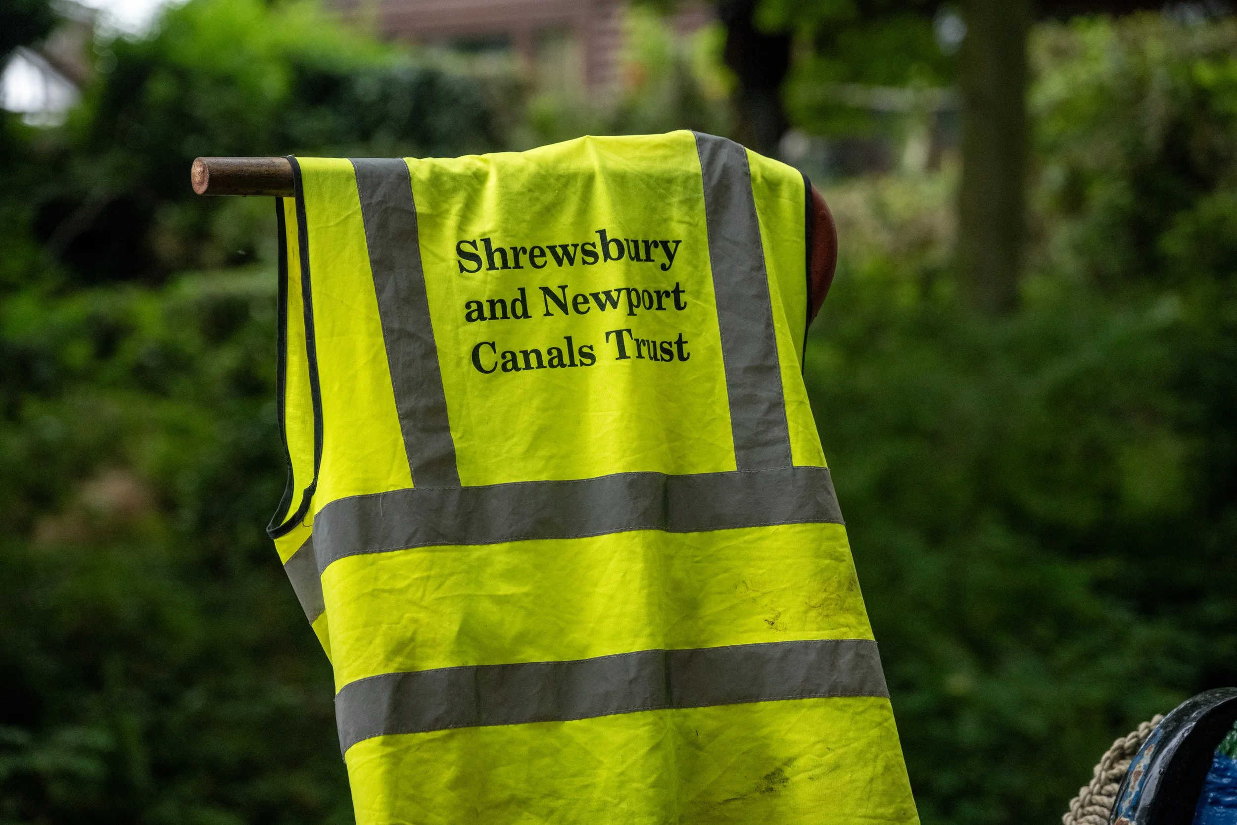 High-visibility yellow vest with black text that reads "Shrewsbury and Newport Canals Trust" hanging on a horizontal wooden rod with a background of green foliage.