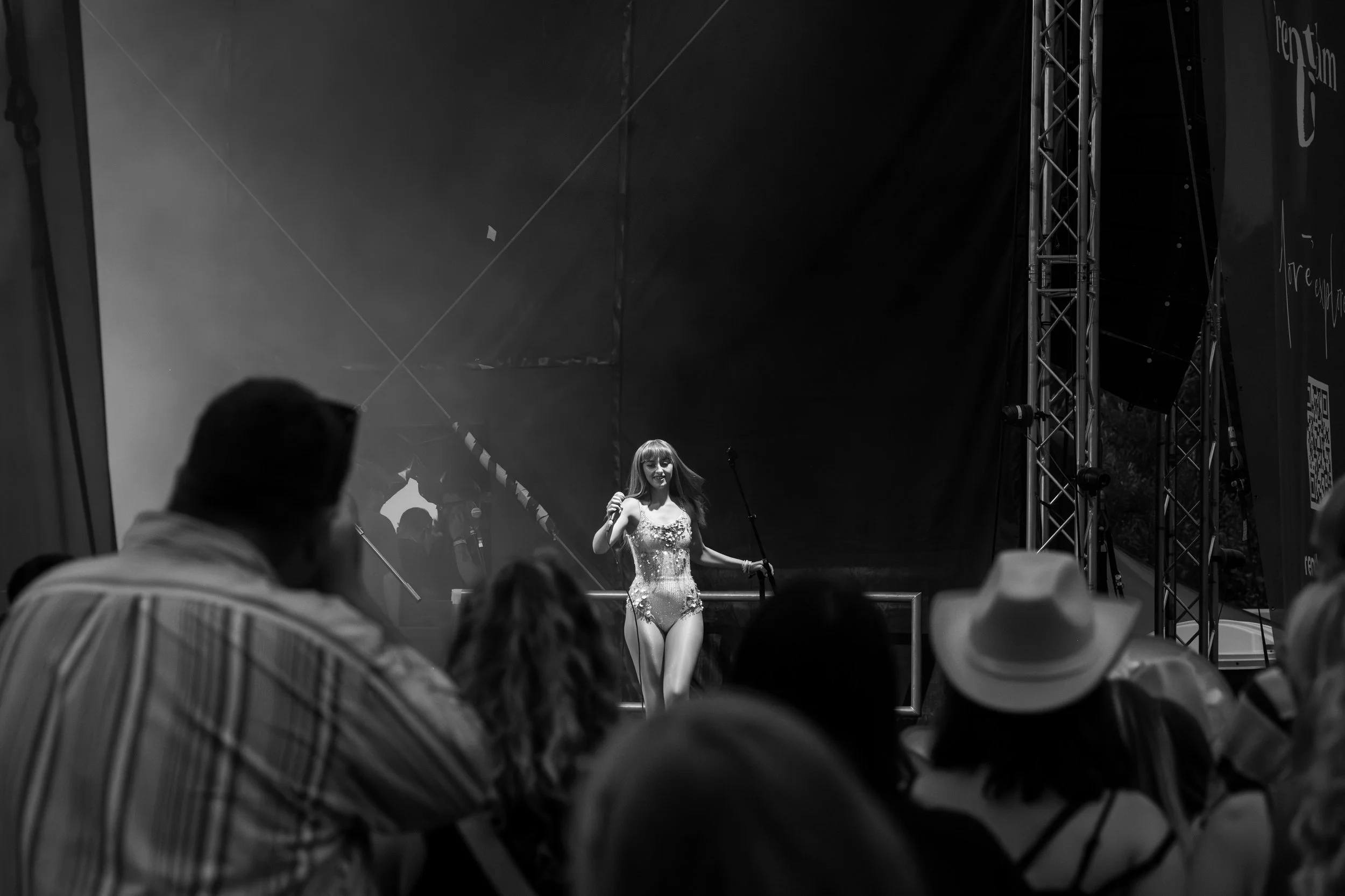 A woman in a fancy, sleeveless dress on stage at a performance or event, holding a microphone, with an audience watching.