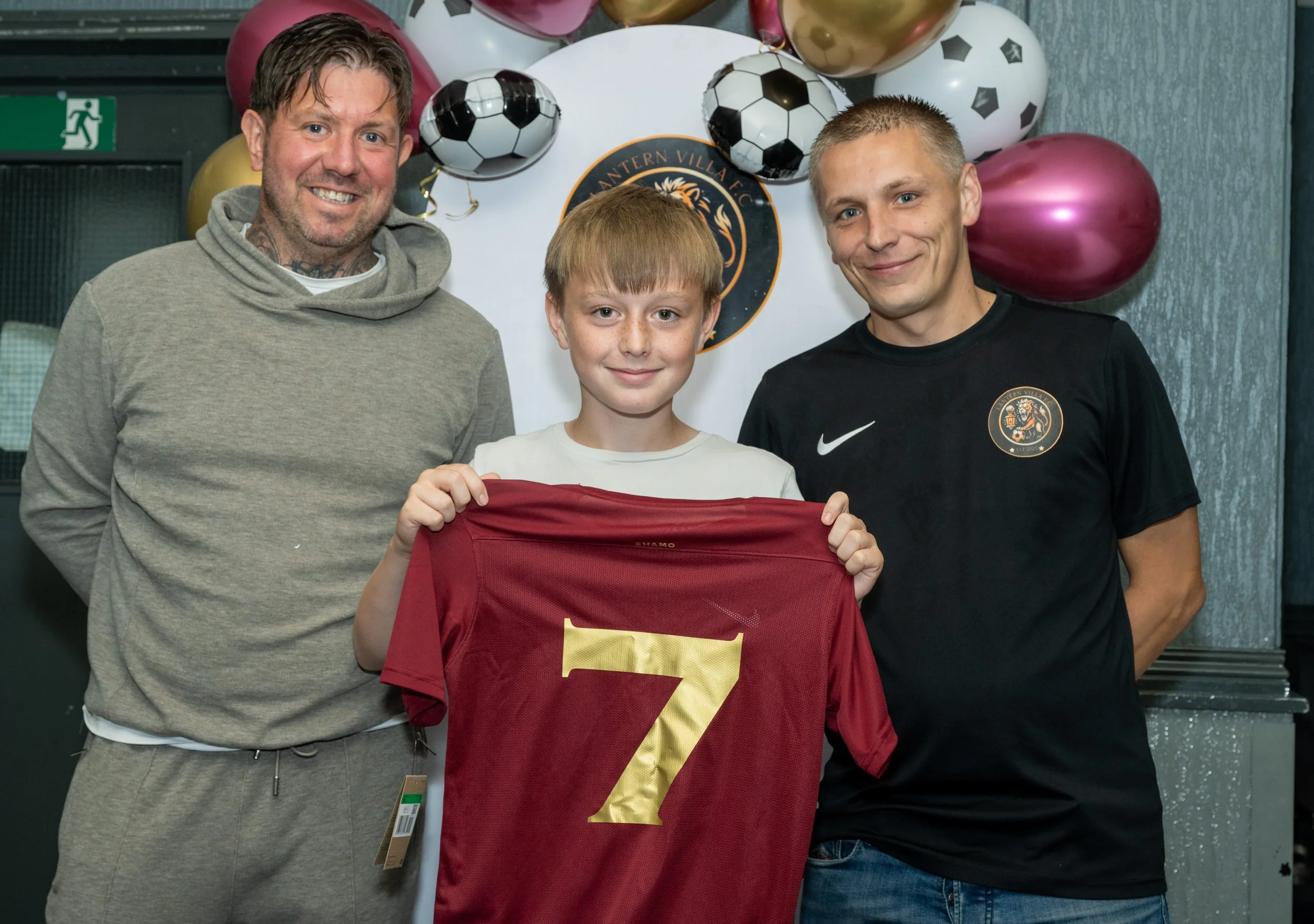 A young boy holding a maroon soccer jersey with the number 7 in gold, standing between two smiling men in front of soccer-themed balloons and banners.