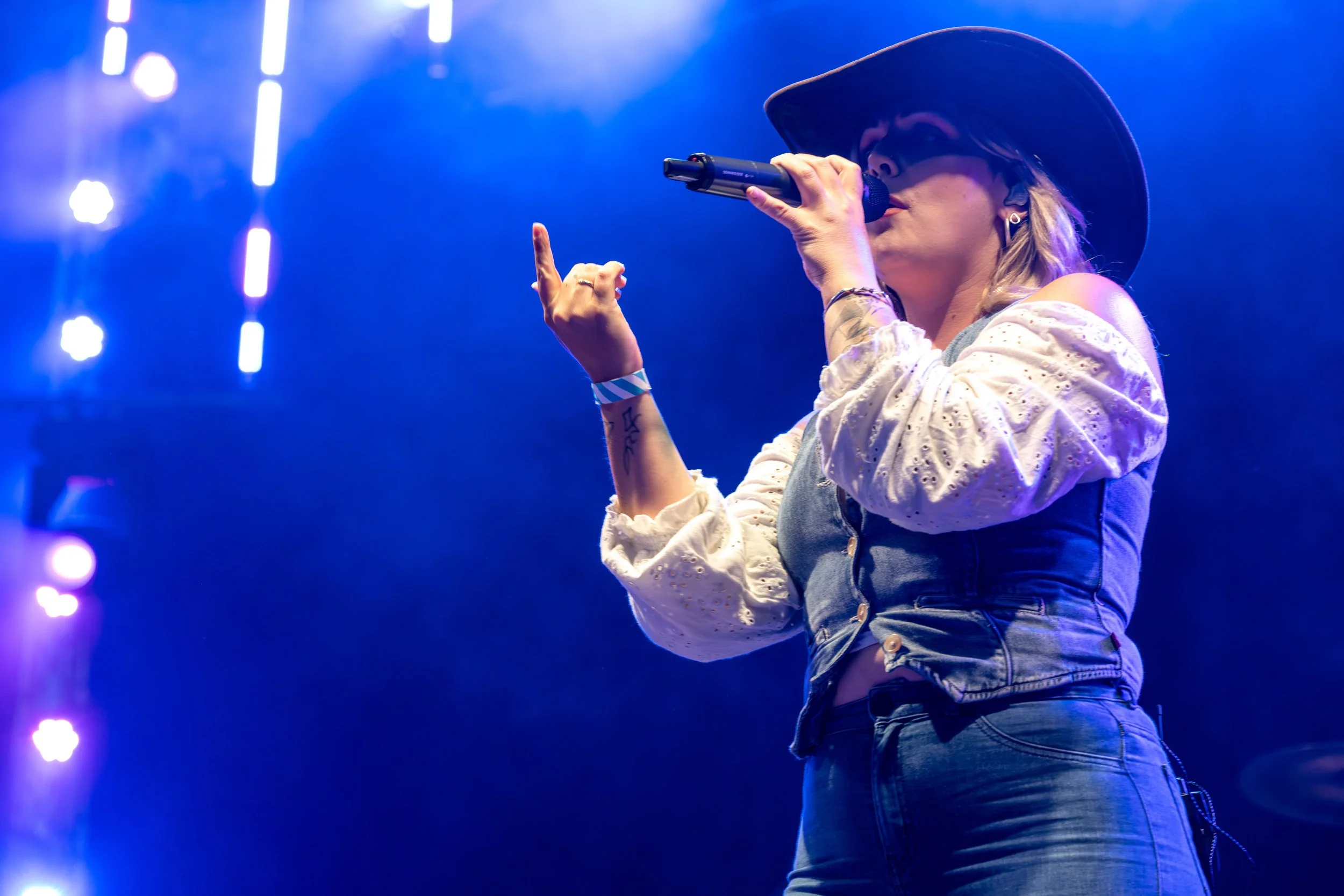 A woman wearing a black hat, denim vest, and jeans singing into a microphone on stage with purple and white stage lights.