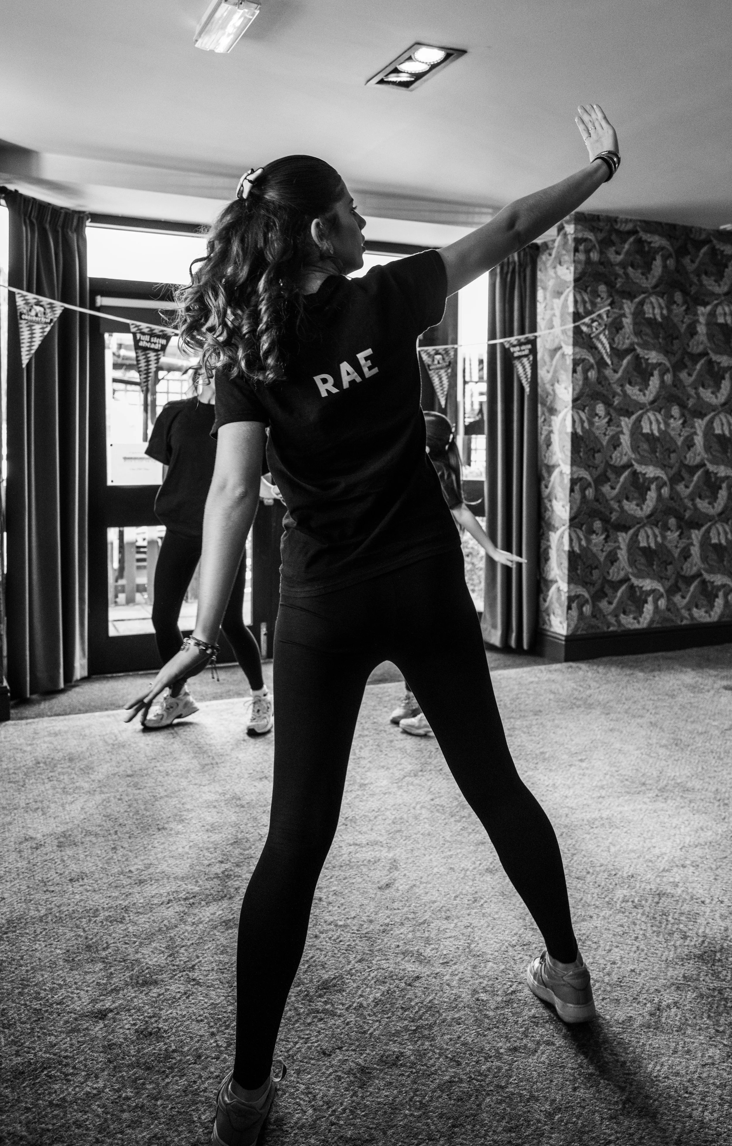A woman with long curly hair, wearing a black shirt with 'RACE' written on the back, is dancing or practicing a dance move indoors. Two other women are visible in the background, also participating in the activity. The room has large windows, curtain