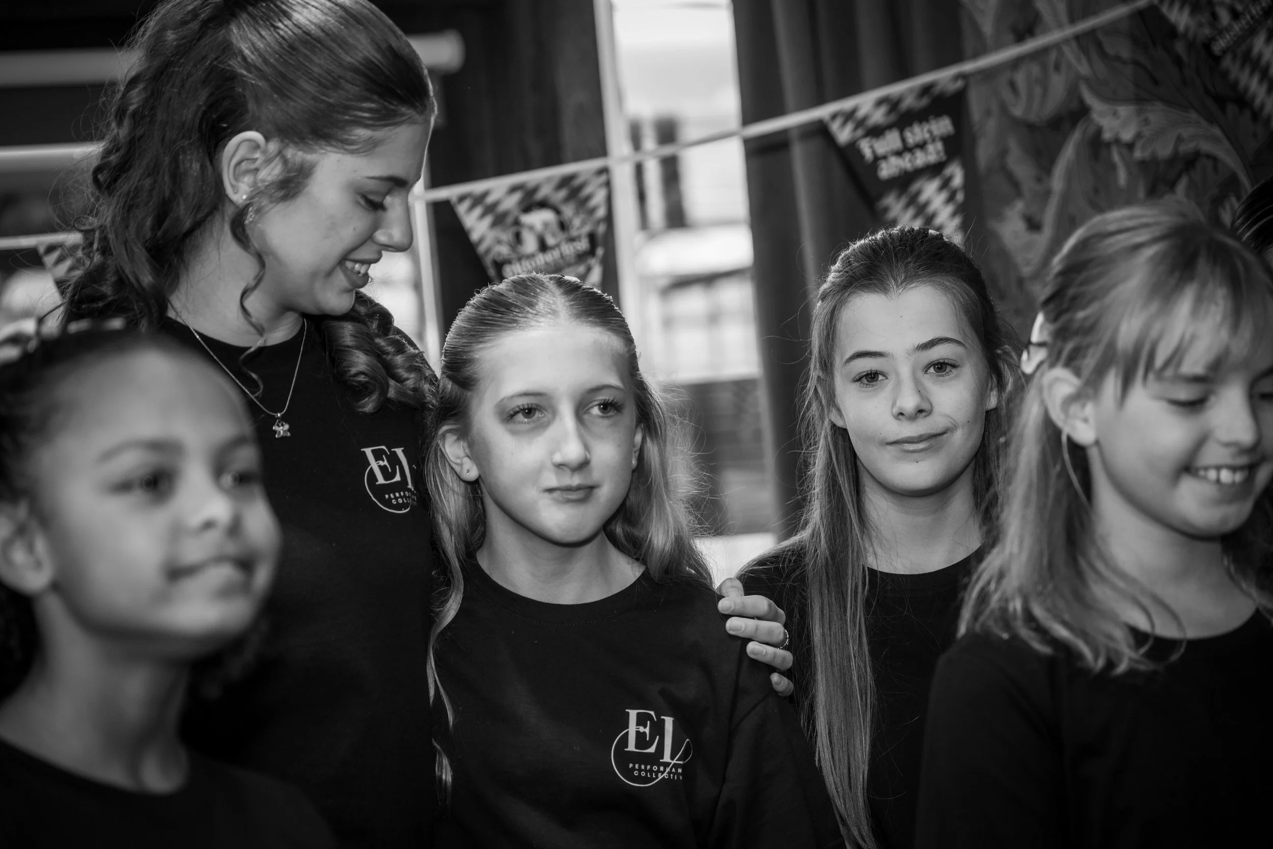 A group of young girls standing together, some smiling, in an indoor setting with banners in the background.