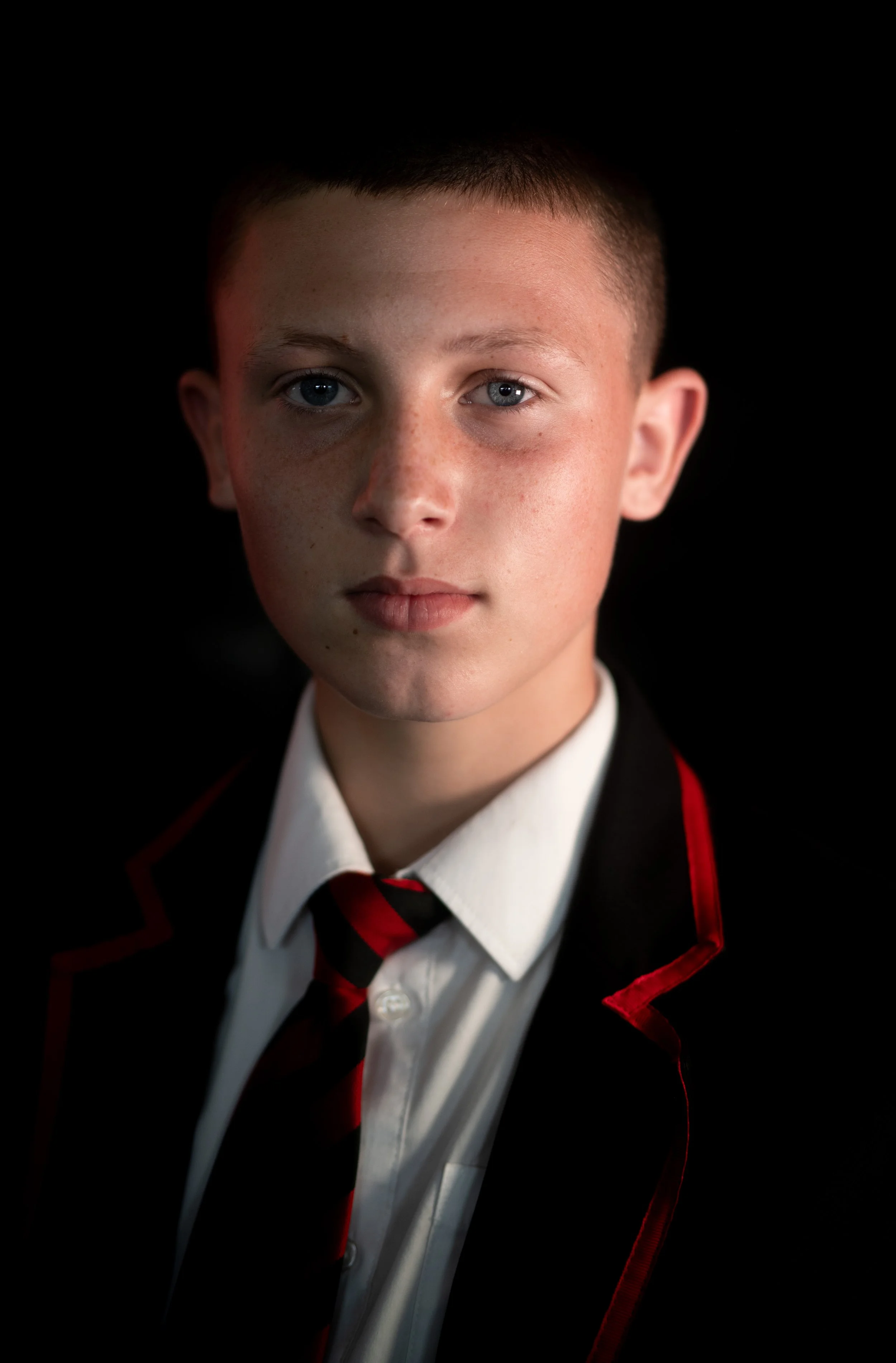 Close-up portrait of a young boy with short brown hair, wearing a white shirt, black blazer with red trim, and a red and black striped tie, against a dark background.