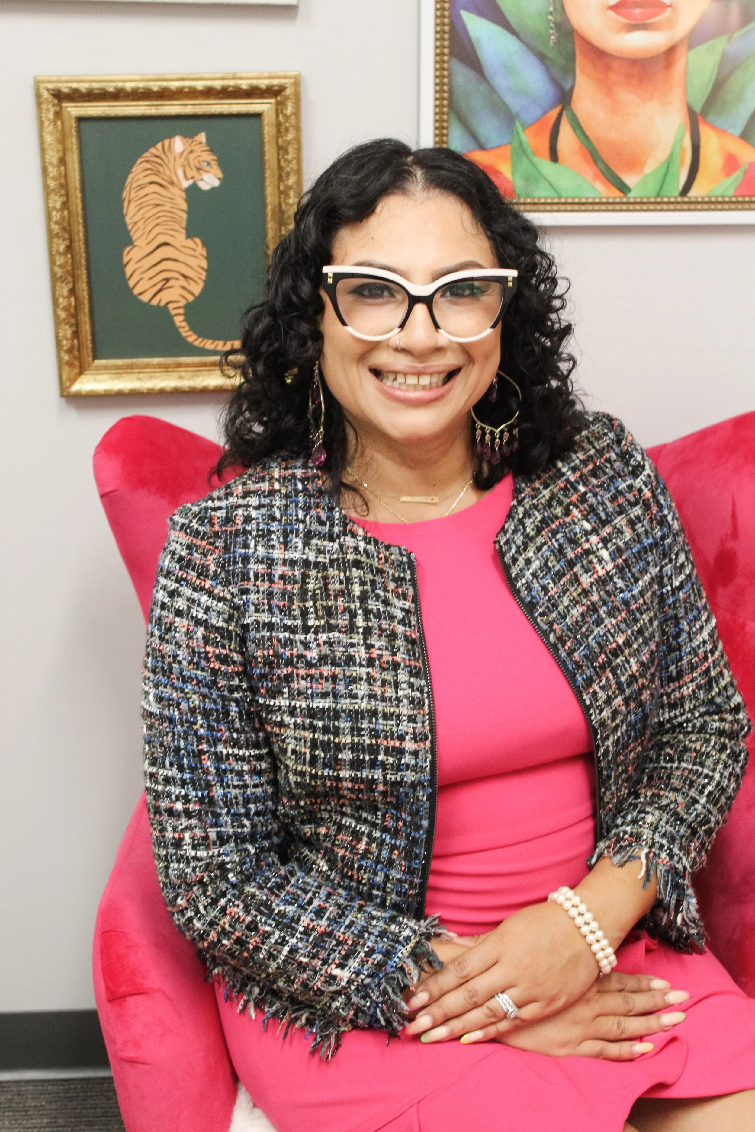 A woman smiling, sitting in a pink chair with portraits hanging behind her. She is wearing glasses, a colorful tweed blazer, a pink dress, pearl jewelry, and has dark curly hair.