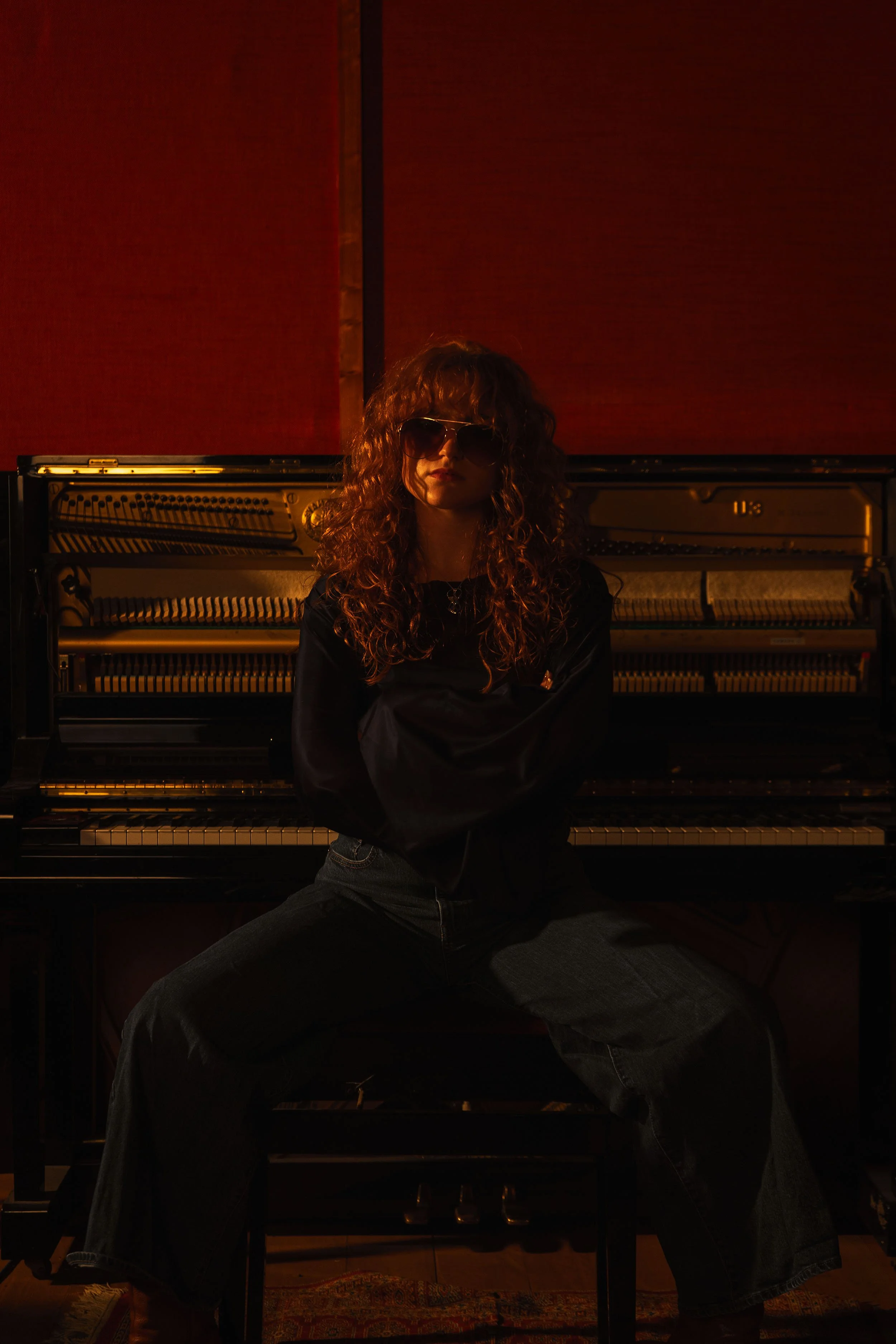 Chloë Wilson with curly hair wearing sunglasses, black top, and jeans sitting on a piano bench in front of an open piano with a dark red background.