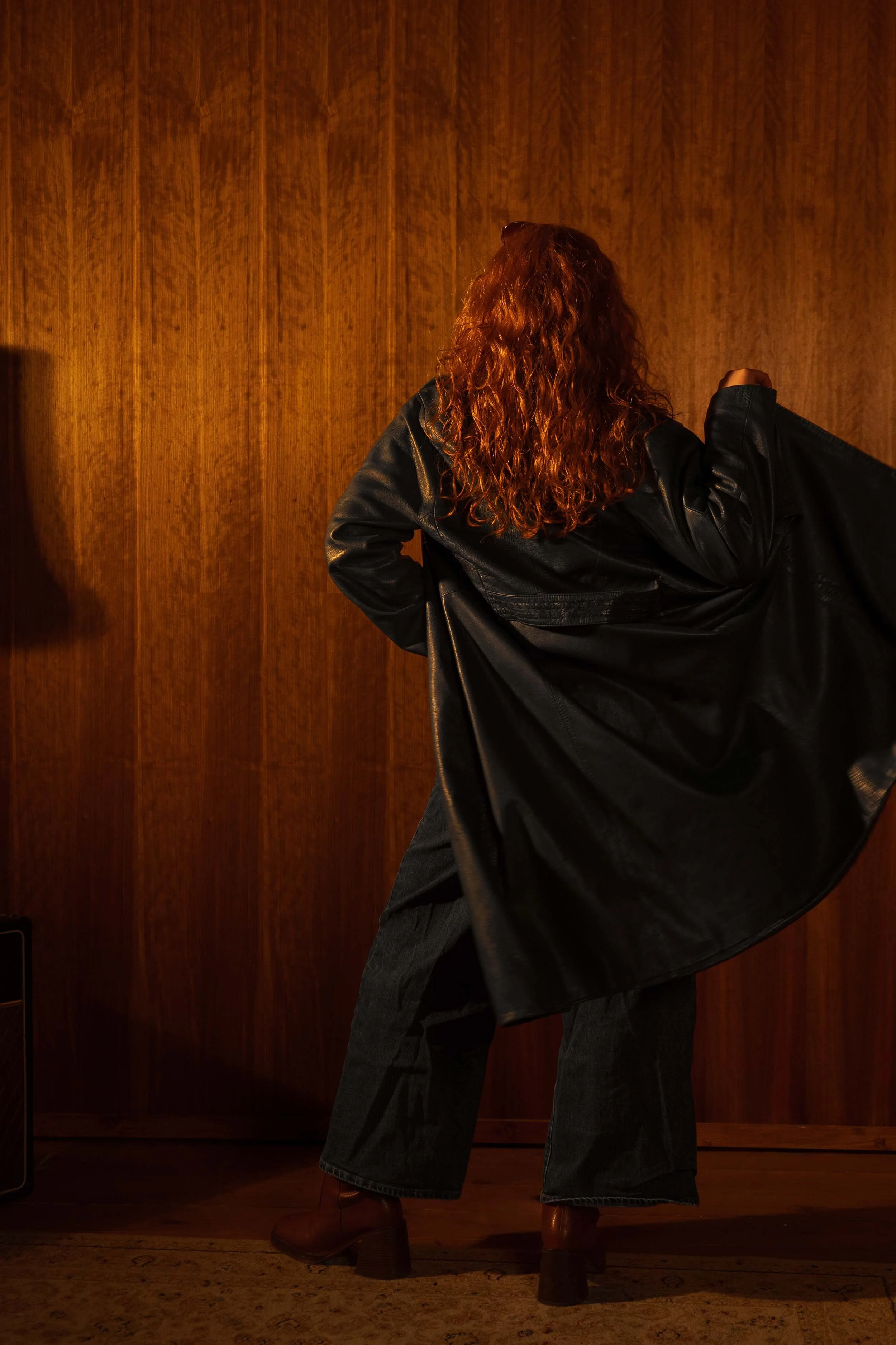 A woman (Chloë Wilson) with curly red hair wearing a black leather jacket, dark jeans, and brown heeled boots, standing in front of a wooden wall, partly turned away from the camera, with one hand holding the jacket open.