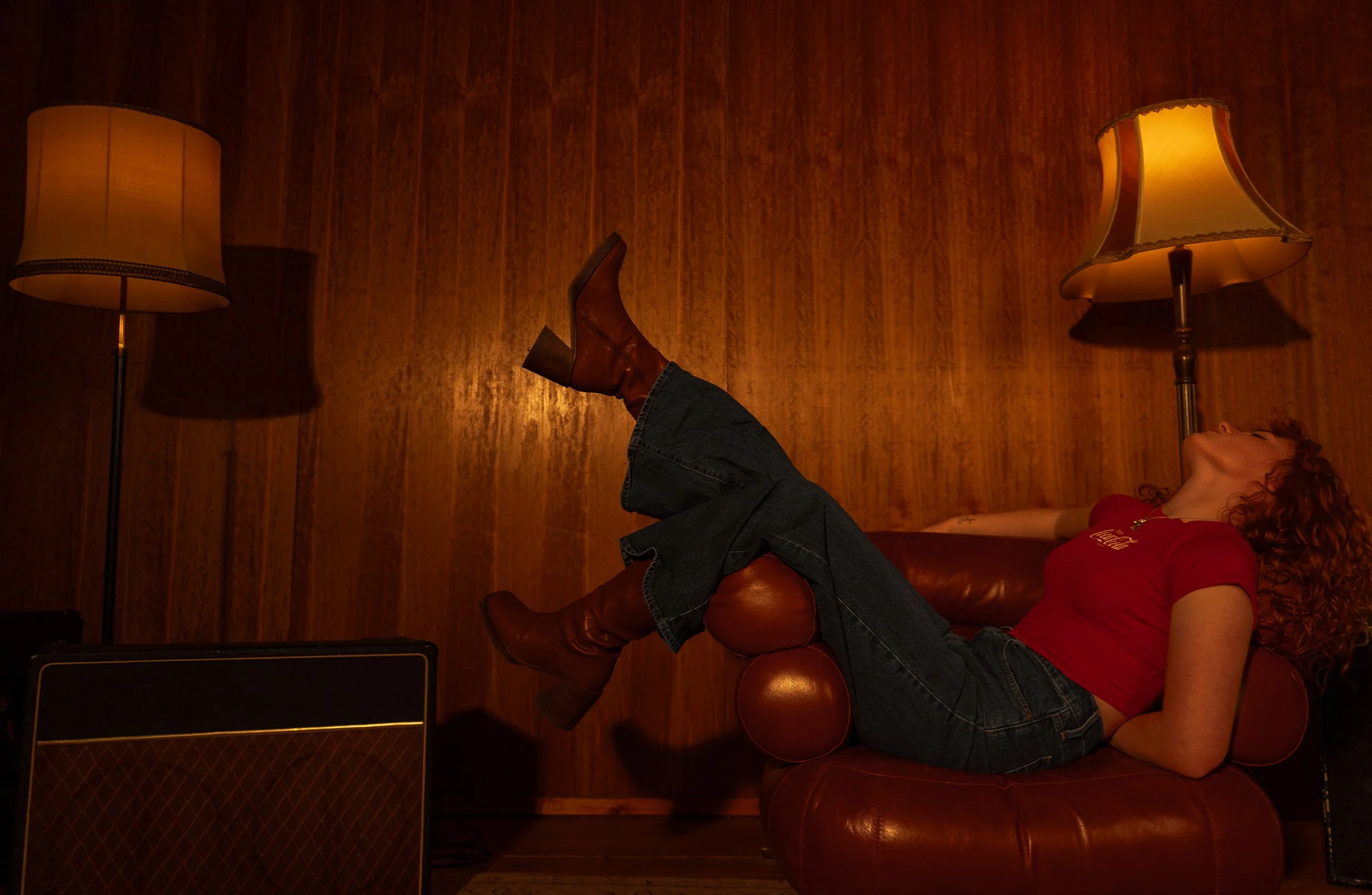 Chloë Wilson with curly red hair lying on a leather couch, wearing a red shirt and boots, with her legs raised and crossed, in a dimly lit room with wooden walls and two floor lamps.
