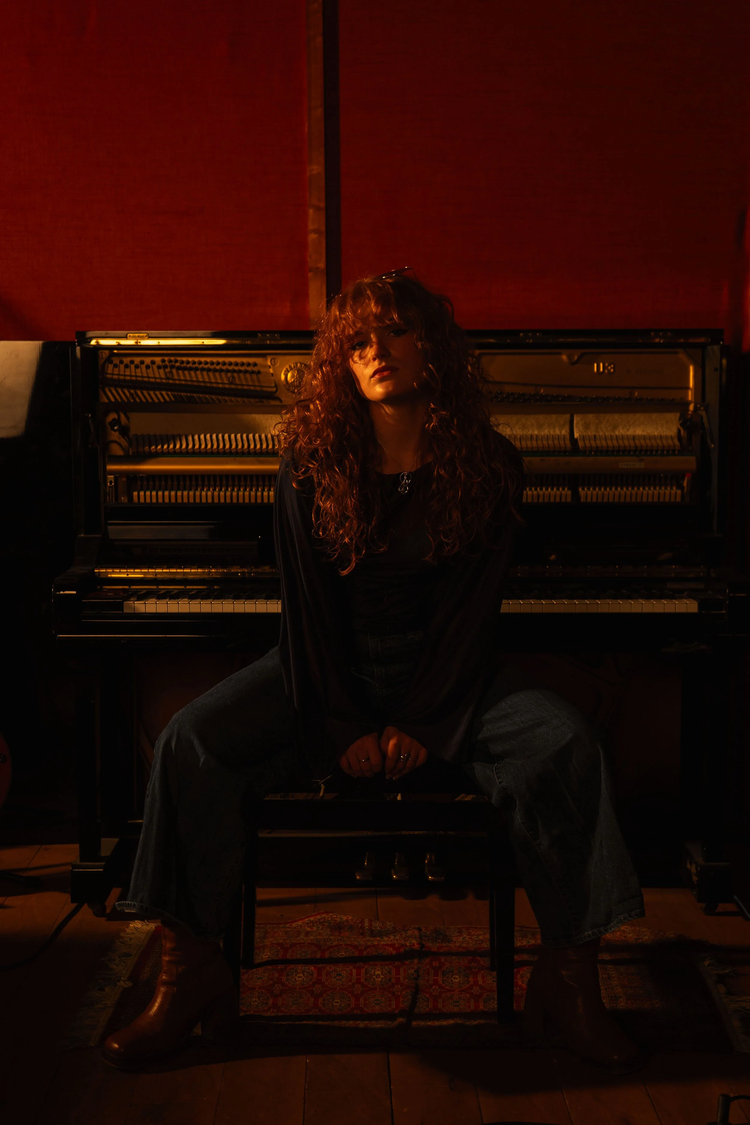 A woman (Chloë Wilson) with curly ginger hair sitting on an open piano bench in front of an upright piano in a dimly lit room with red wall panels.
