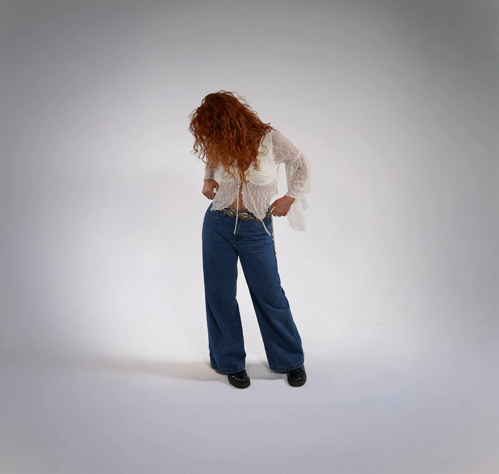Chloë Wilson with long curly red hair looking down, wearing a white lace top and blue jeans, standing in a plain white studio background.