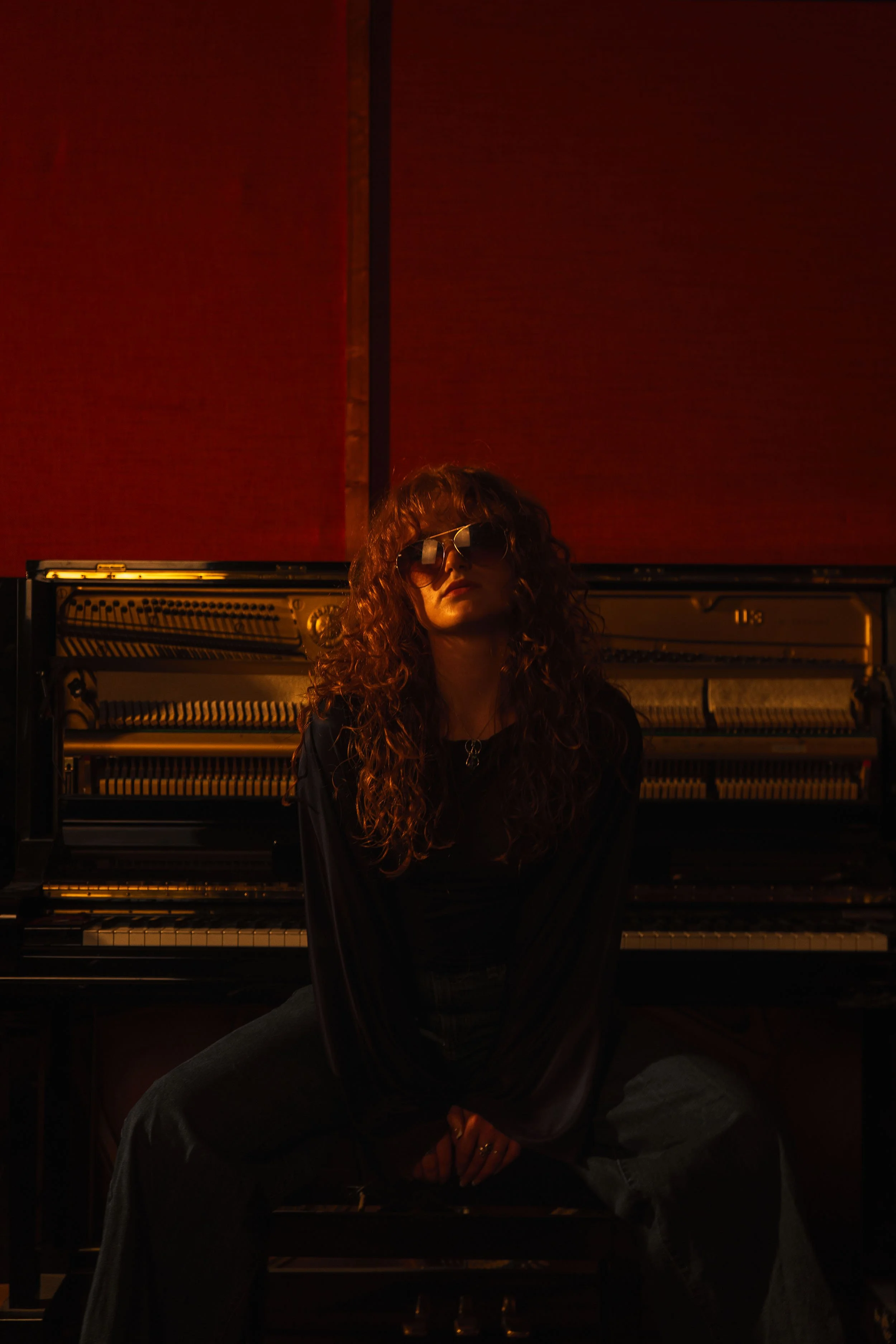 Chloë Wilson with curly hair wearing sunglasses, sitting in a dimly lit room in front of an old piano with exposed strings and hammers.