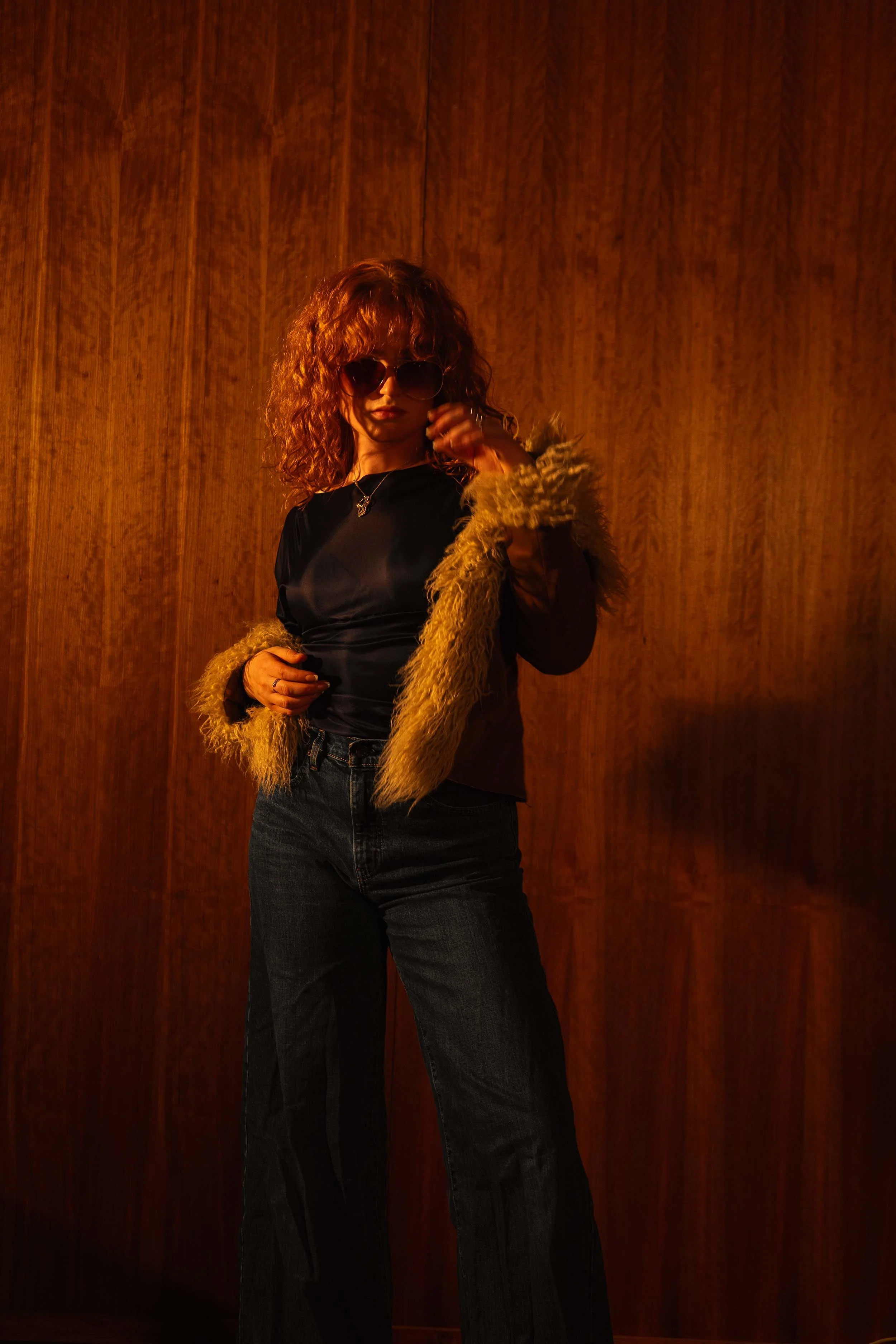 Chloë Wilson with curly ginger hair wearing sunglasses, a black top, a furry coat, and dark blue jeans, standing against a wooden wall background.