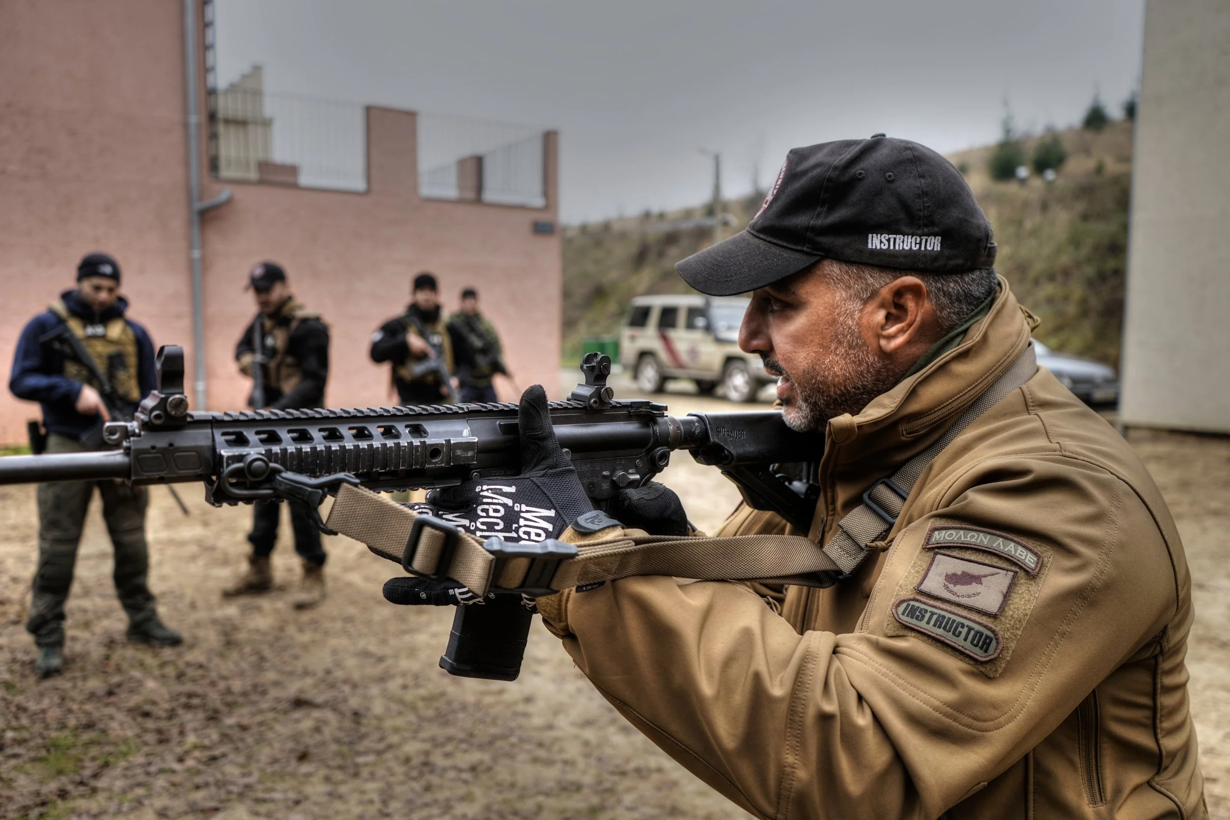 Tactical shooting instructor in a coyote jacket, aiming rifle with a group of armed individuals in the background. Teaching courses, firearms training, close protection training, and military contractor training.