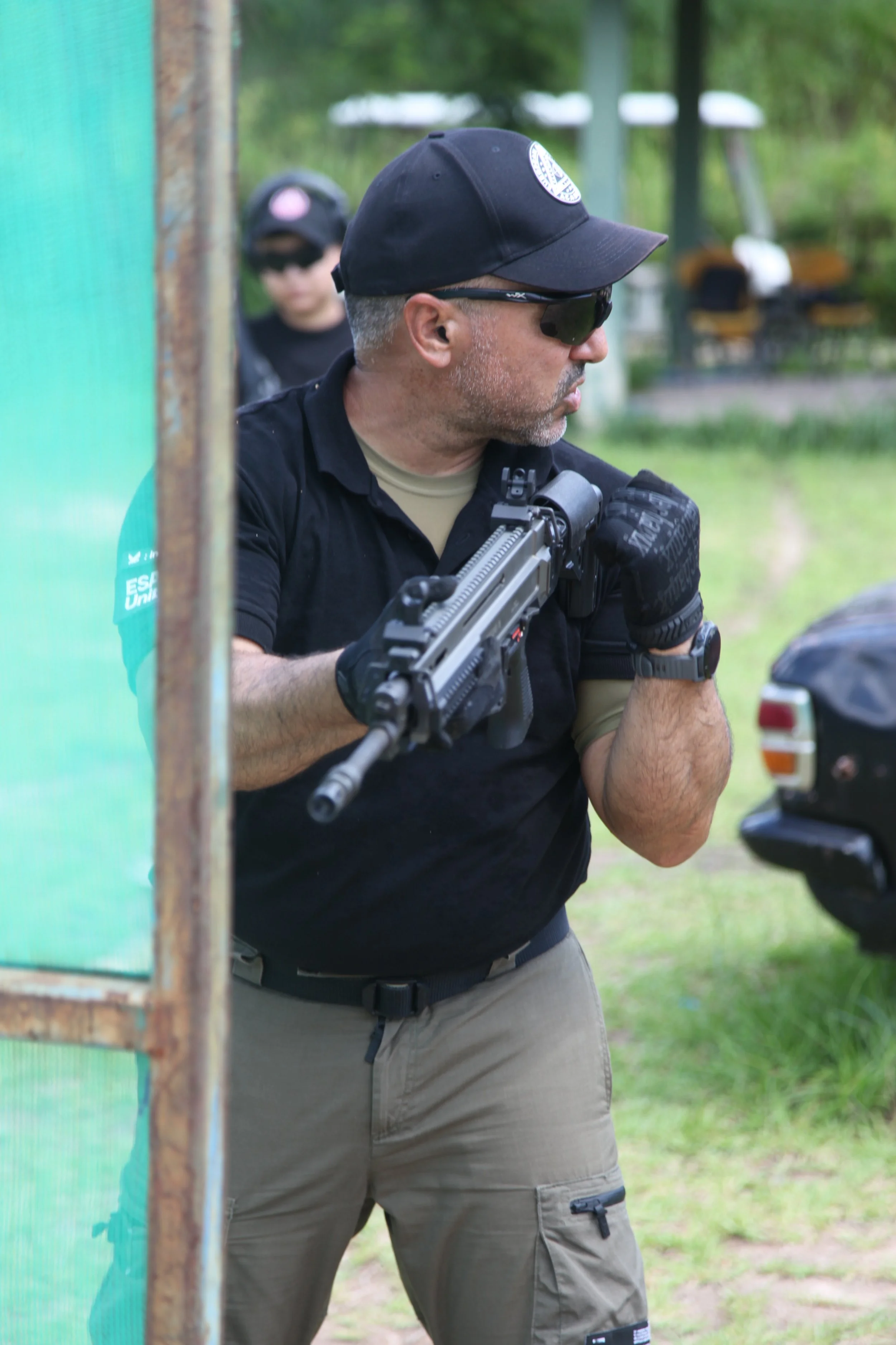 A man wearing sunglasses, a black cap, black shirt, and khaki cargo pants is holding a gun at an outdoor shooting range.