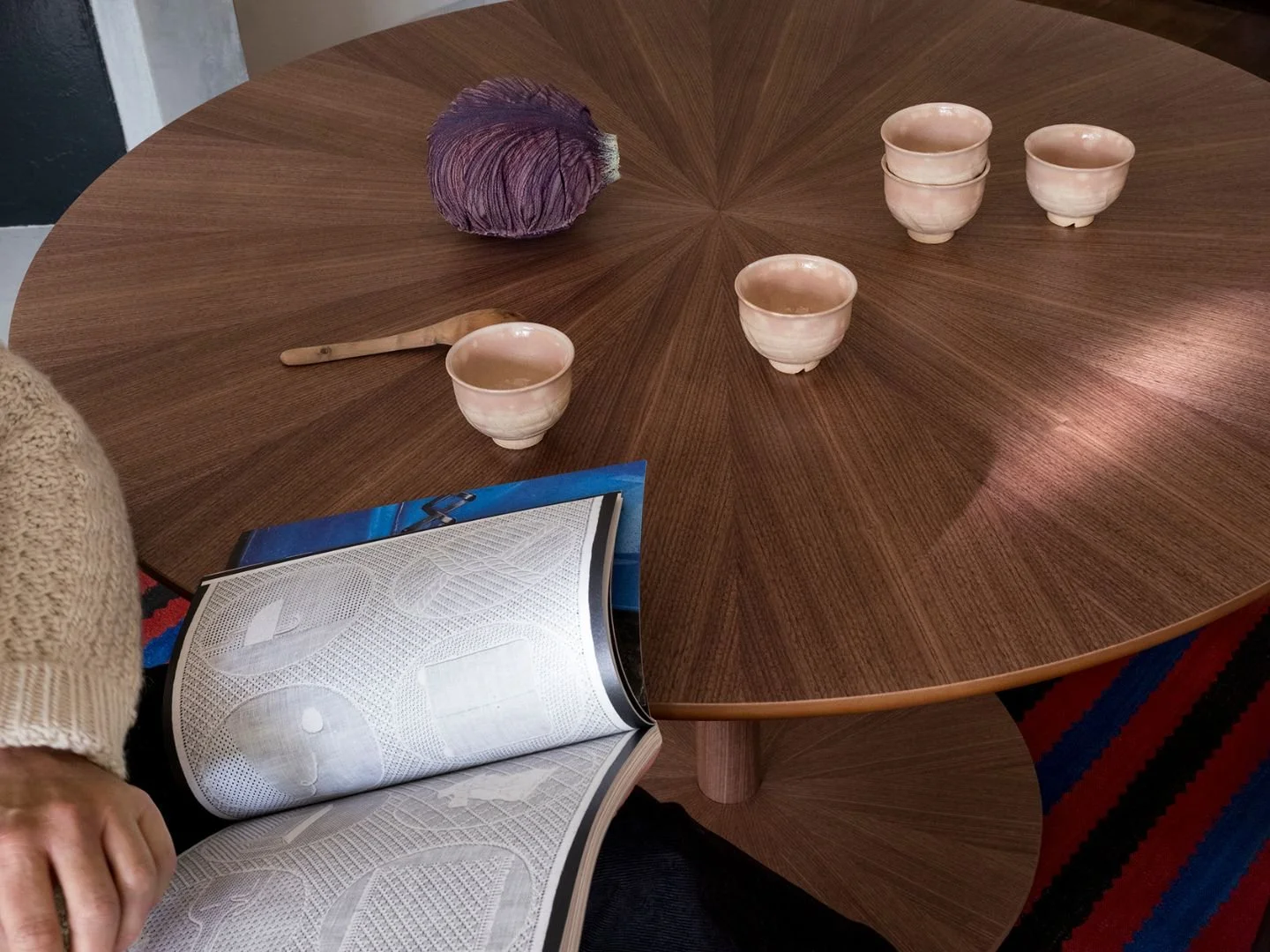 Ein runder Holztisch mit vier rosa Teetassen, einem violetten, strukturierten Gegenstand, einem Holzspatel und einer aufgeschlagenen Zeitschrift. Links ist ein Teil eines Arms zu sehen, der einen beigen Pullover trägt.