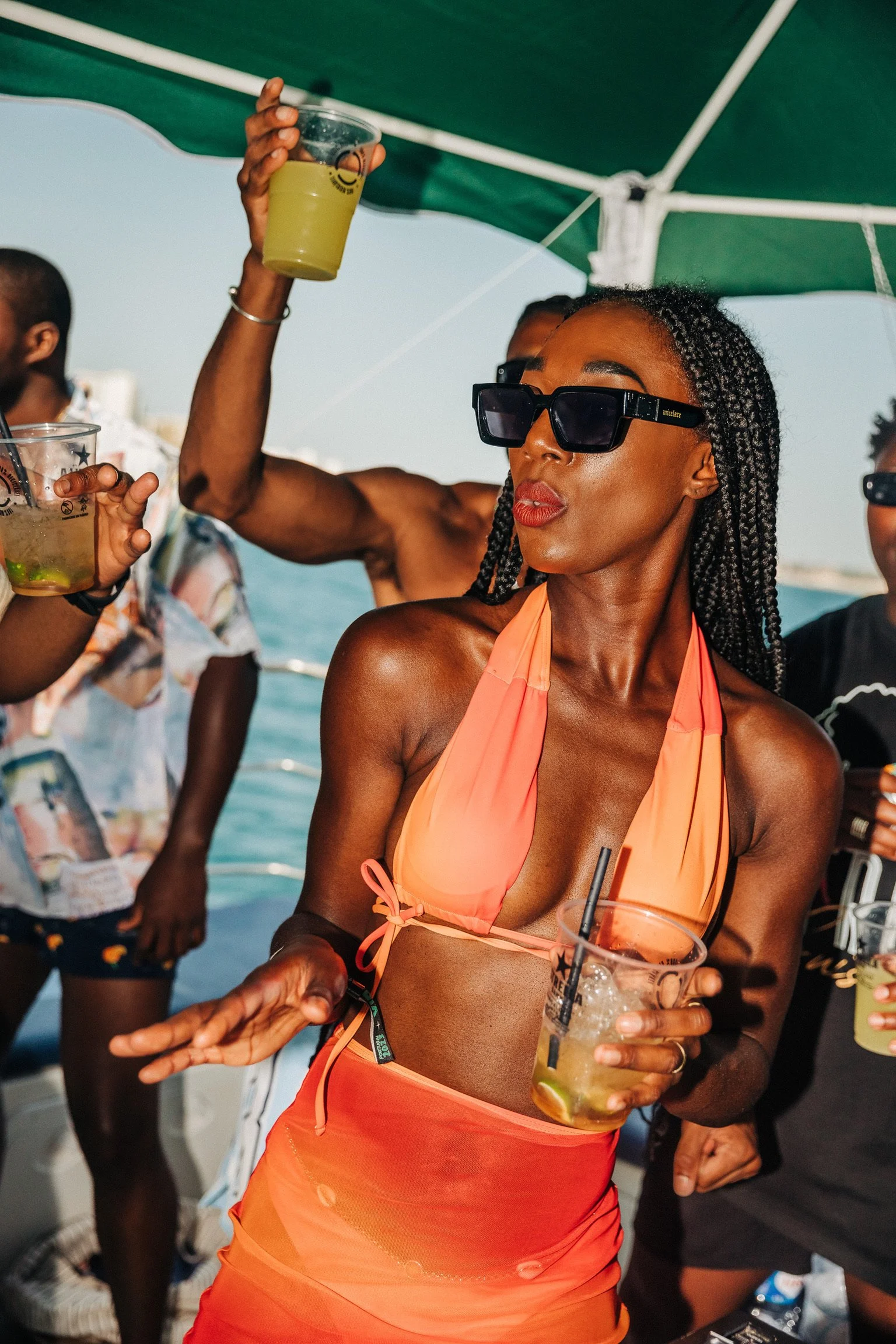 A woman wearing sunglasses, an orange bikini top, and a matching skirt enjoying a drink on a boat during daytime with other people around, some holding drinks and a green canopy overhead.