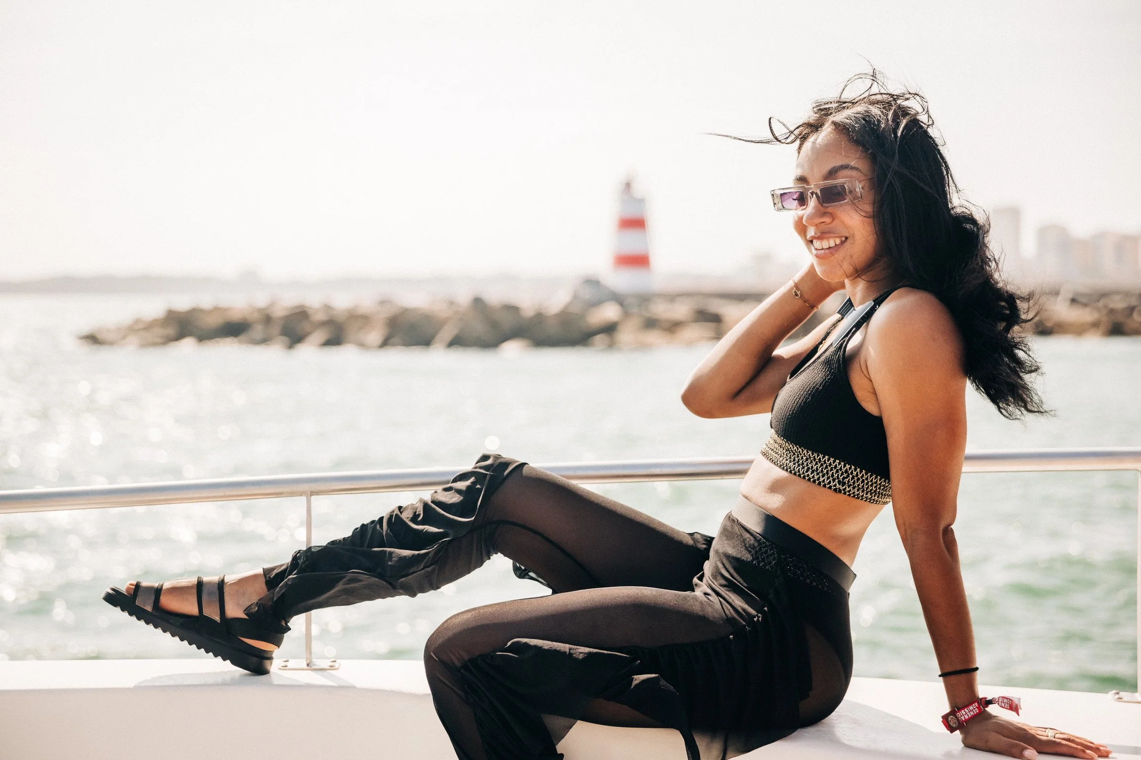 Smiling woman with black hair wearing sunglasses and athletic clothing sitting on a boat with water and a lighthouse in the background.