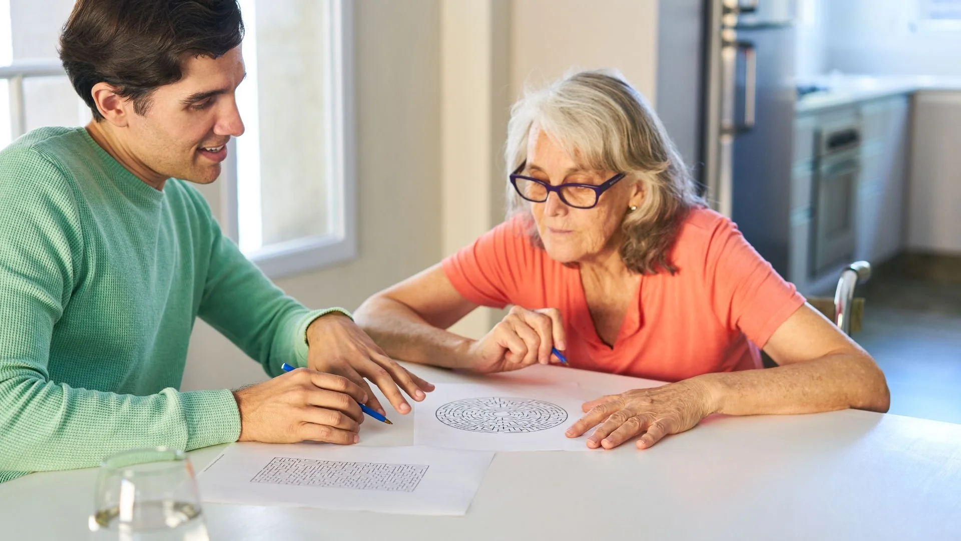 A caregiver in a green sweater engaging in a cognitive activity with an elderly woman wearing glasses and an orange shirt at a table.