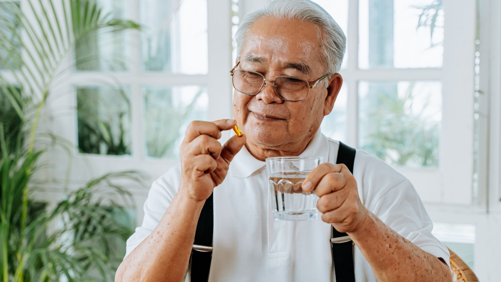 An elderly Asian man seated by a window, holding a medication capsule and a glass of water, preparing to take his medicine.