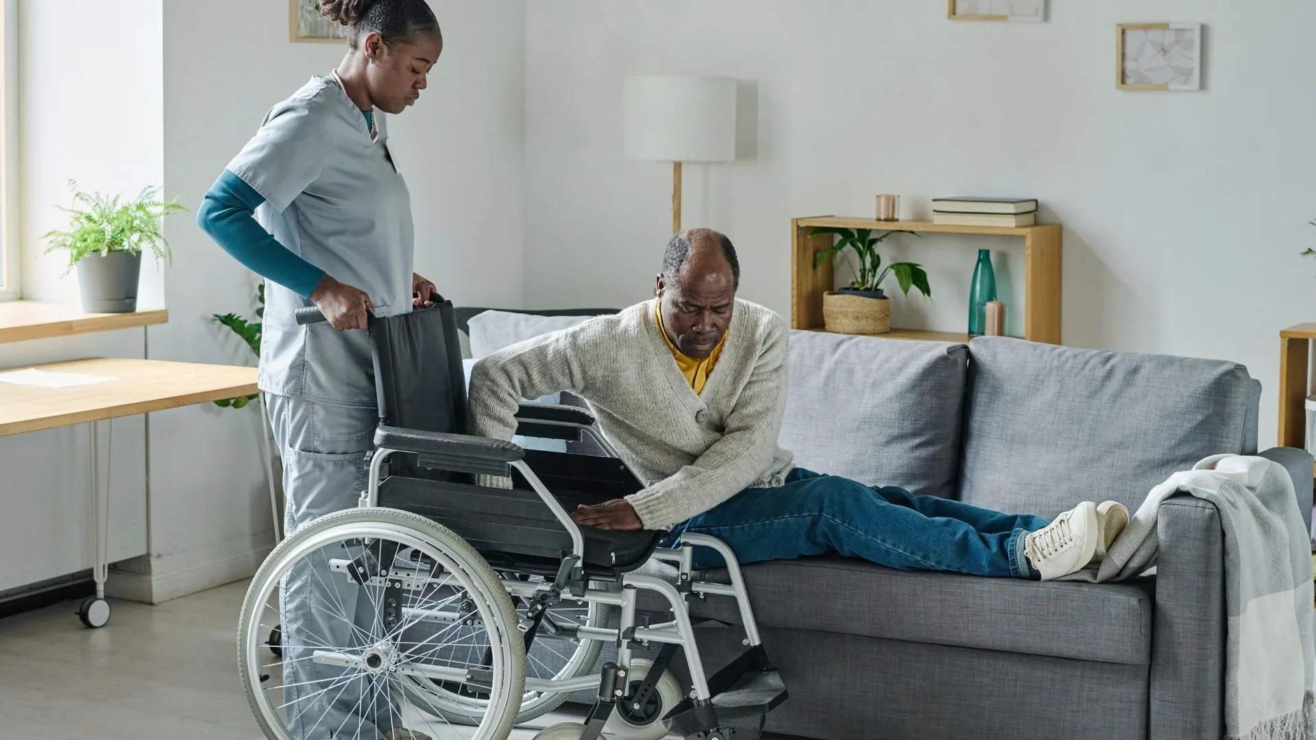 A caregiver assisting an elderly African American man in transitioning from a sofa to a wheelchair in a modern living room.