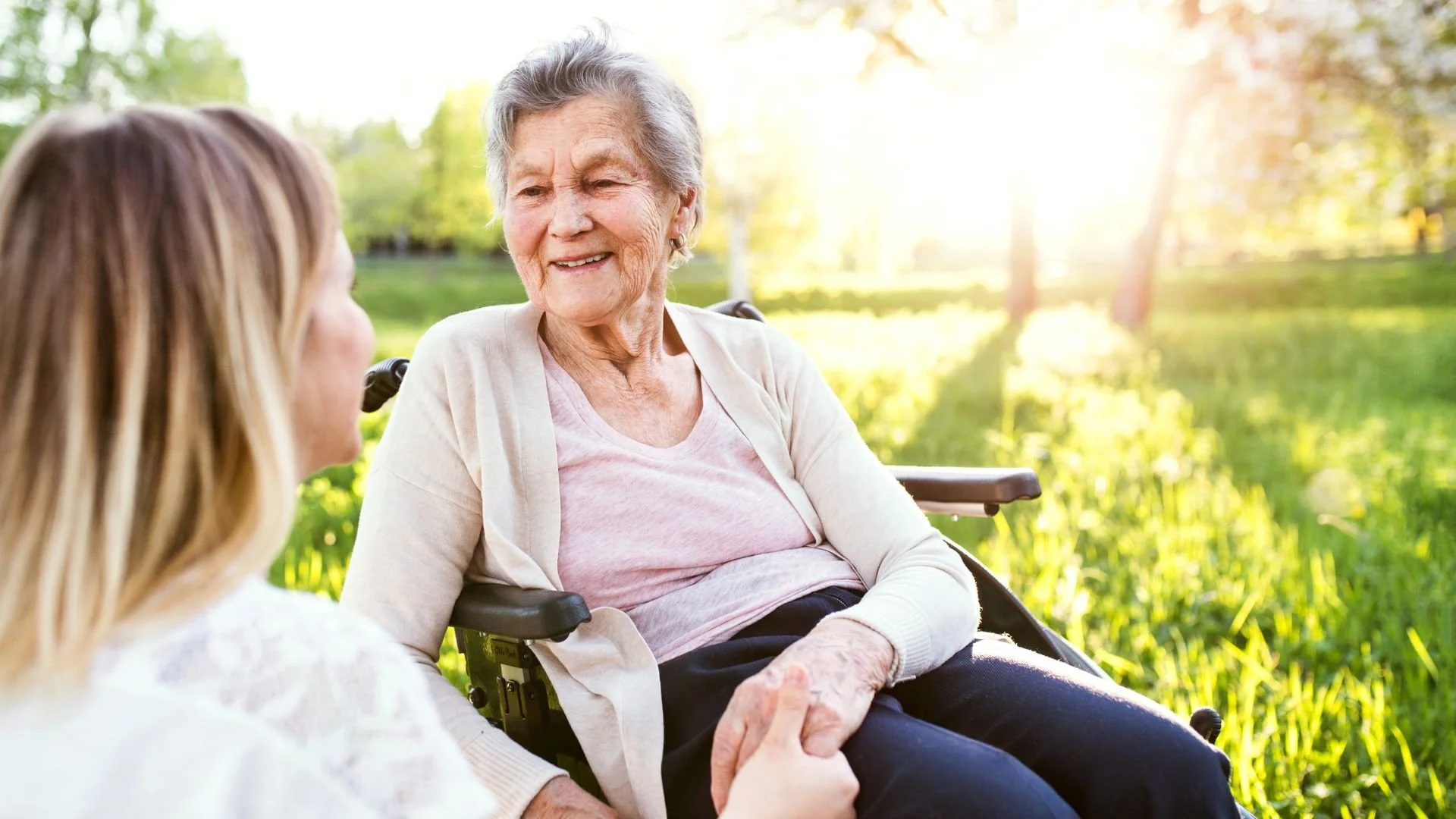 A caregiver holds the hand of an elderly person seated in a wheelchair, providing comfort and companionship during a peaceful outdoor stroll.