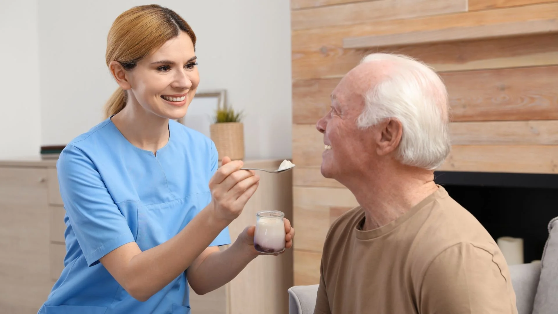 A smiling caregiver feeding yogurt to an elderly bald man in a casual home environment, highlighting assisted feeding services.