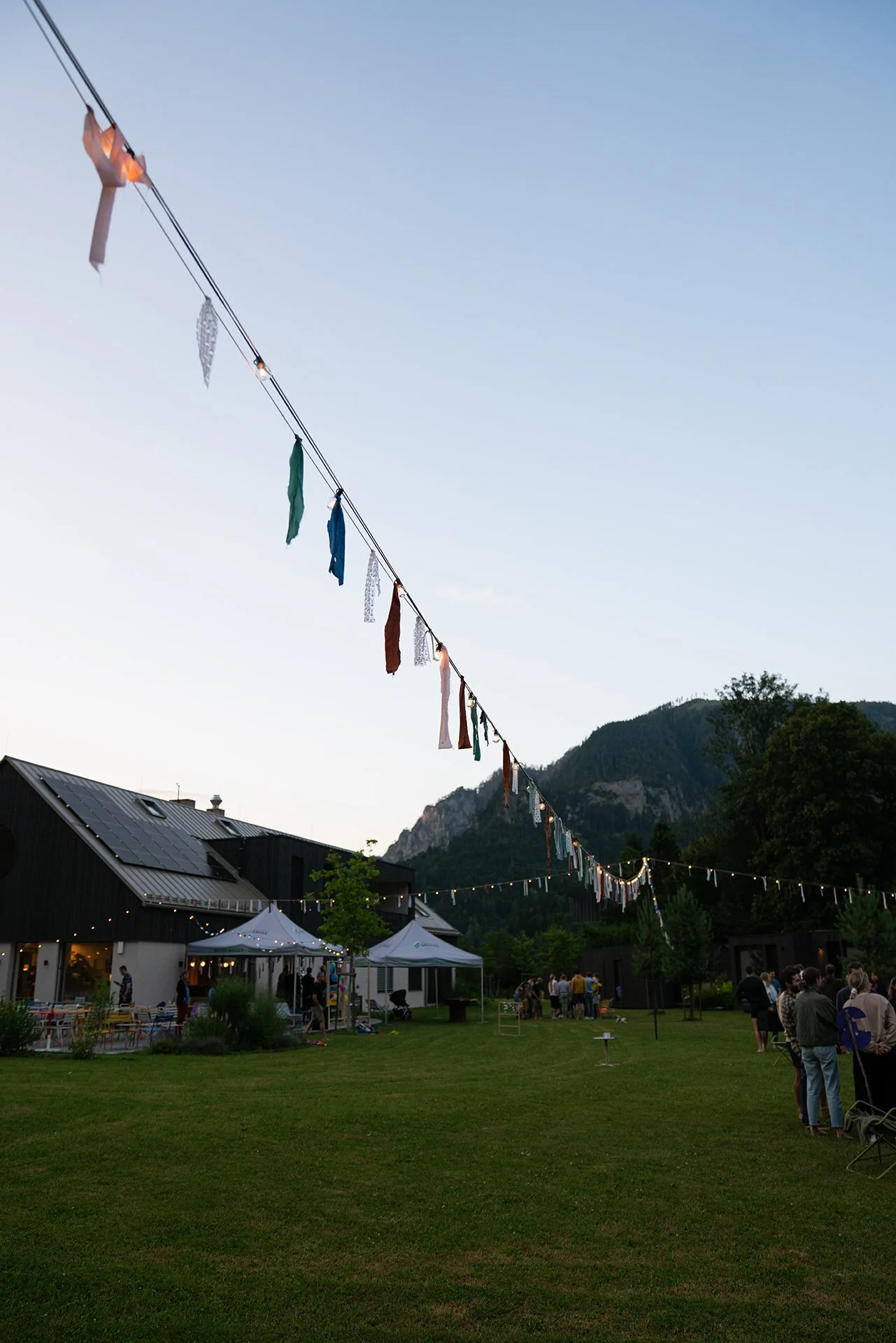 Ein Fest im Freien bei Sonnenuntergang mit bunten Wimpeln an einer Schnur, Tischen, Zelten und einer Gruppe von Menschen im Garten, umgeben von Bergen und Gebäuden.