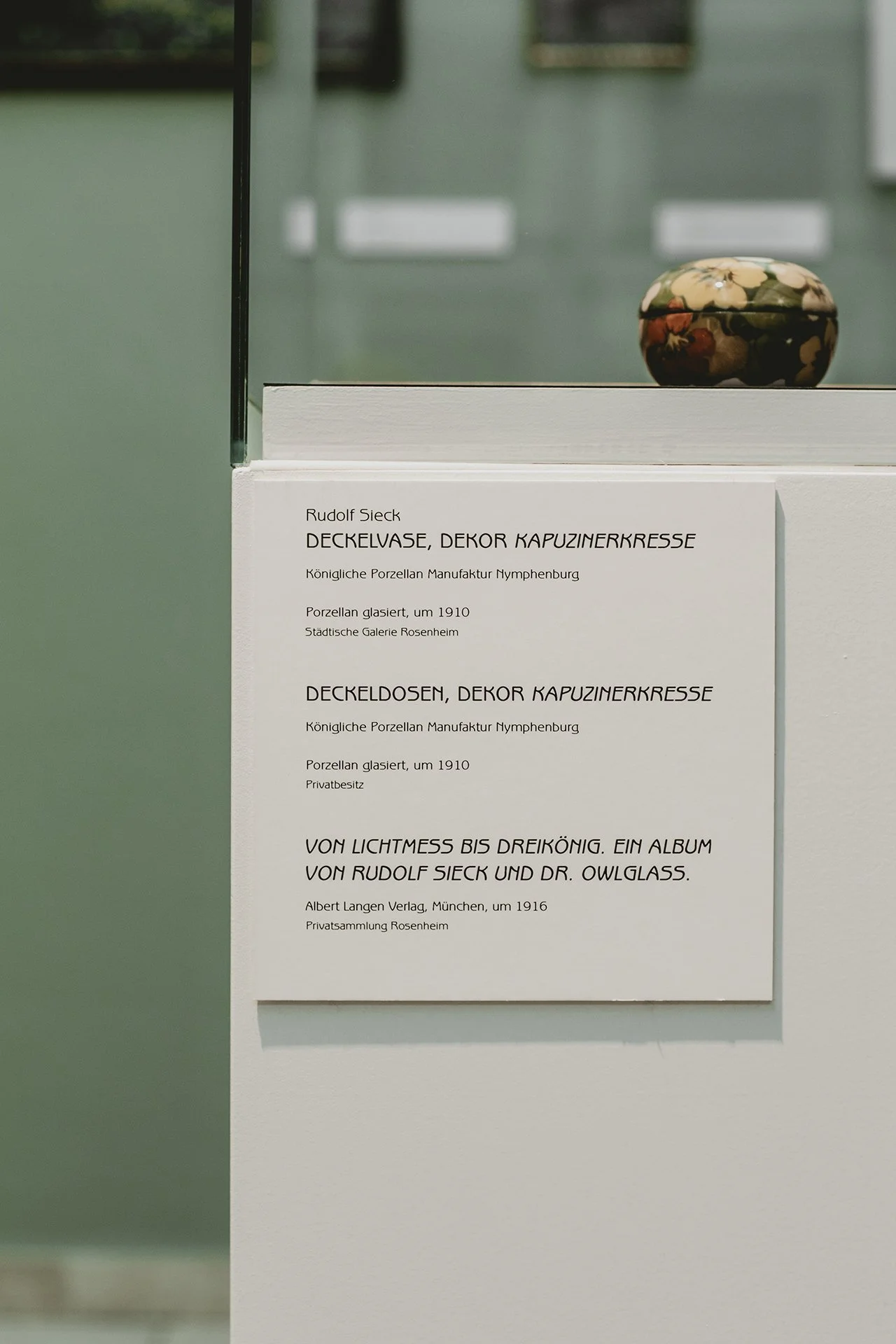 Ein kleines, bemaltes Porzellanrequisit auf einem weißen Sockel, mit einem Ausstellungsschild darunter, das auf eine Ausstellung im Museum Rosenheim hinweist.