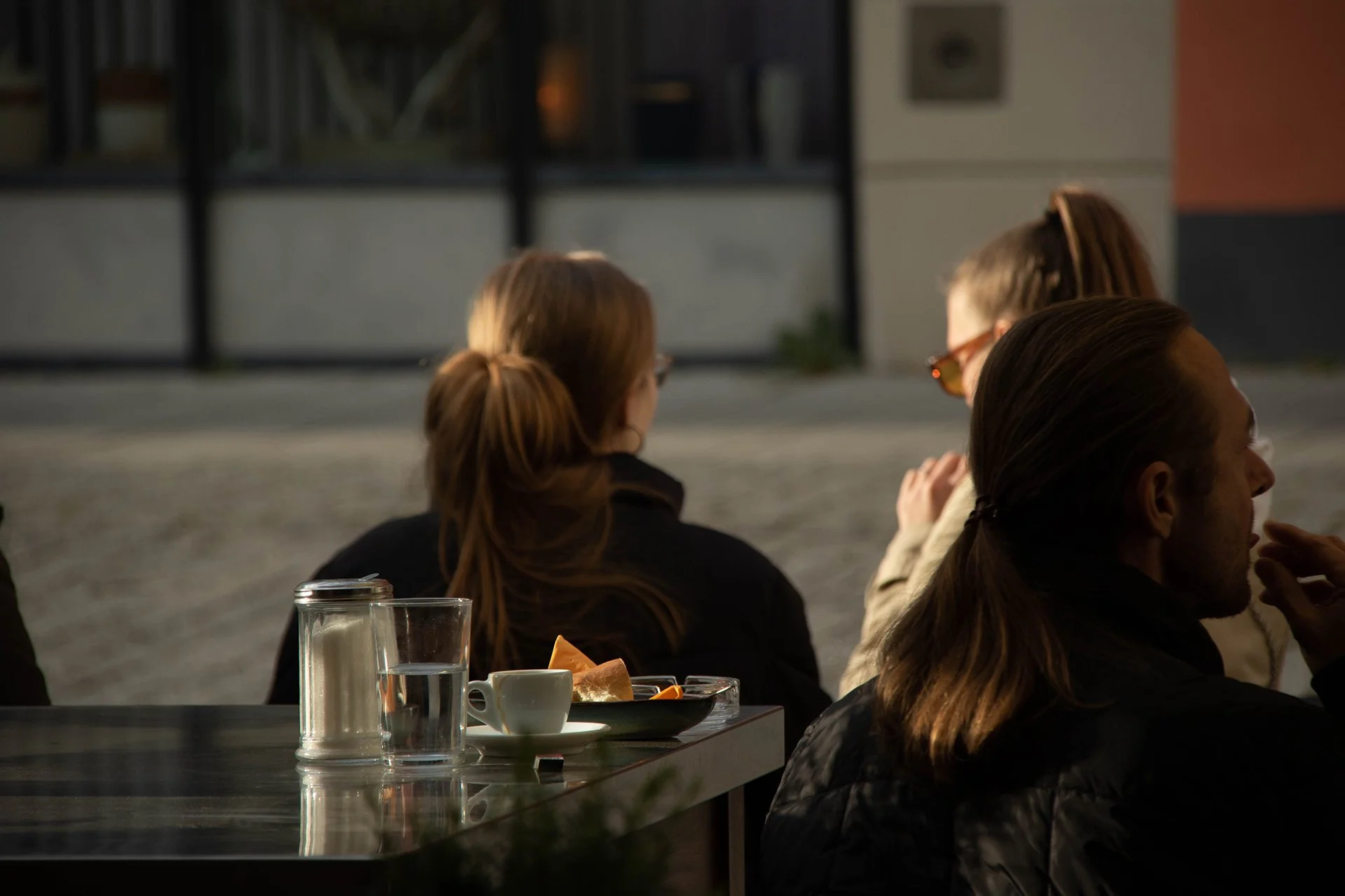 Menschen sitzen an einem Tisch in einem Café im Freien, bei Sonnenuntergang, mit Mahlzeiten und Getränken. Im Hintergrund eine Haustür mit Glaselementen.